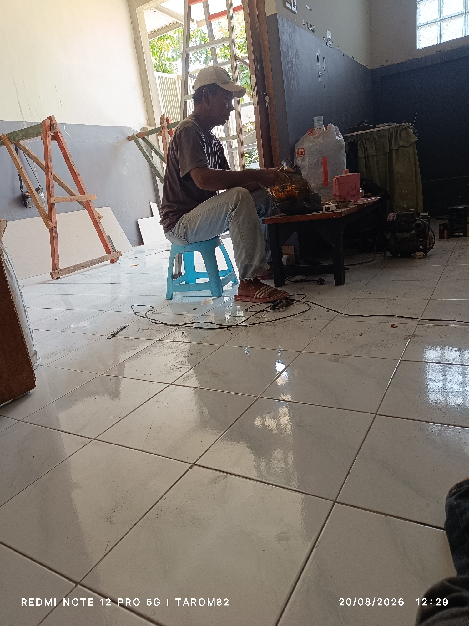 In Sruni, Indonesia, a man wearing a brown t-shirt, blue jeans, and a white cap is seated on a blue plastic stool. He is on a tiled floor in what appears to be a workshop or storage area, with natural light streaming in from an open doorway. In front of him, on a dark wooden table, are several items including a plastic bag, a small pink container, and a bowl filled with an orange substance, possibly food. He is actively engaged with something in the bowl. The environment is cluttered with construction materials, including wooden scaffolding and large white sheets of what could be drywall, suggesting ongoing renovation or building work. A black electrical cord snakes across the floor from the table towards the man's feet. The scene is dated 20/08/2026 at 12:29.