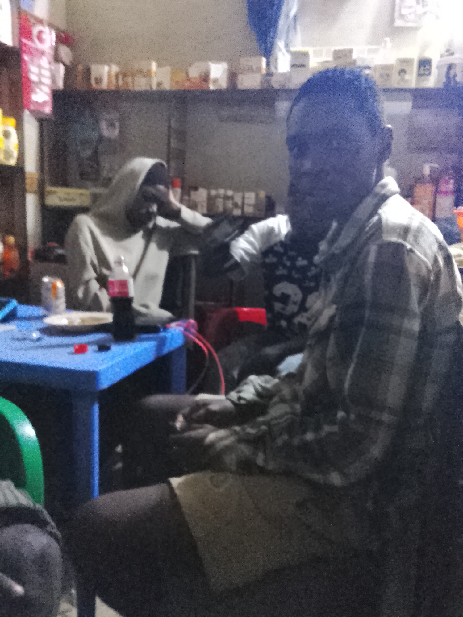 In Durumi, Nigeria, a dimly lit indoor scene captures a moment around a bright blue table. Three individuals are present. In the foreground, a man with dark skin and short hair wears a plaid shirt. To his left, another man, partially obscured by the table, is seated and wearing a hooded garment. Further back, a third person is visible wearing a dark t-shirt with a graphic design. On the blue table, a white plate with food, a can of soda, and a bottle of cola are arranged. Shelves with various items are visible in the background, suggesting a domestic or storage area. The overall lighting is low, with a visible grain effect on the image.