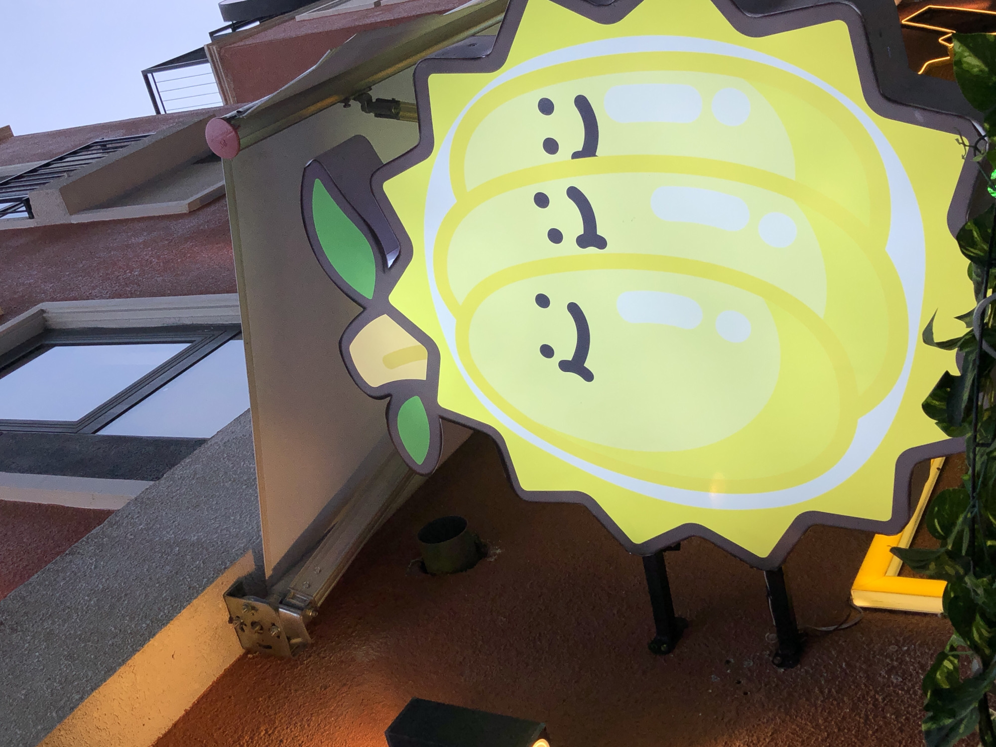 A cartoonish, bright yellow sign depicting three pods of durian fruit is mounted on the exterior of a building. Each durian pod has two black dots for eyes and a curved line for a smile. The sign has a jagged, brown border and is supported by black metal legs. A green leaf shape is attached to the left side of the sign. The building behind the sign has a textured, reddish-brown facade. To the left, a section of a building with a window is visible, along with a metal railing and a staircase. To the right, lush green foliage and a yellow neon light fixture are present. The sky above is overcast and appears to be twilight or early morning. The setting is an urban environment, likely a street-facing establishment in Đà Nẵng, Vietnam, given the context. There is no explicit activity or event taking place; the image captures a static signage.