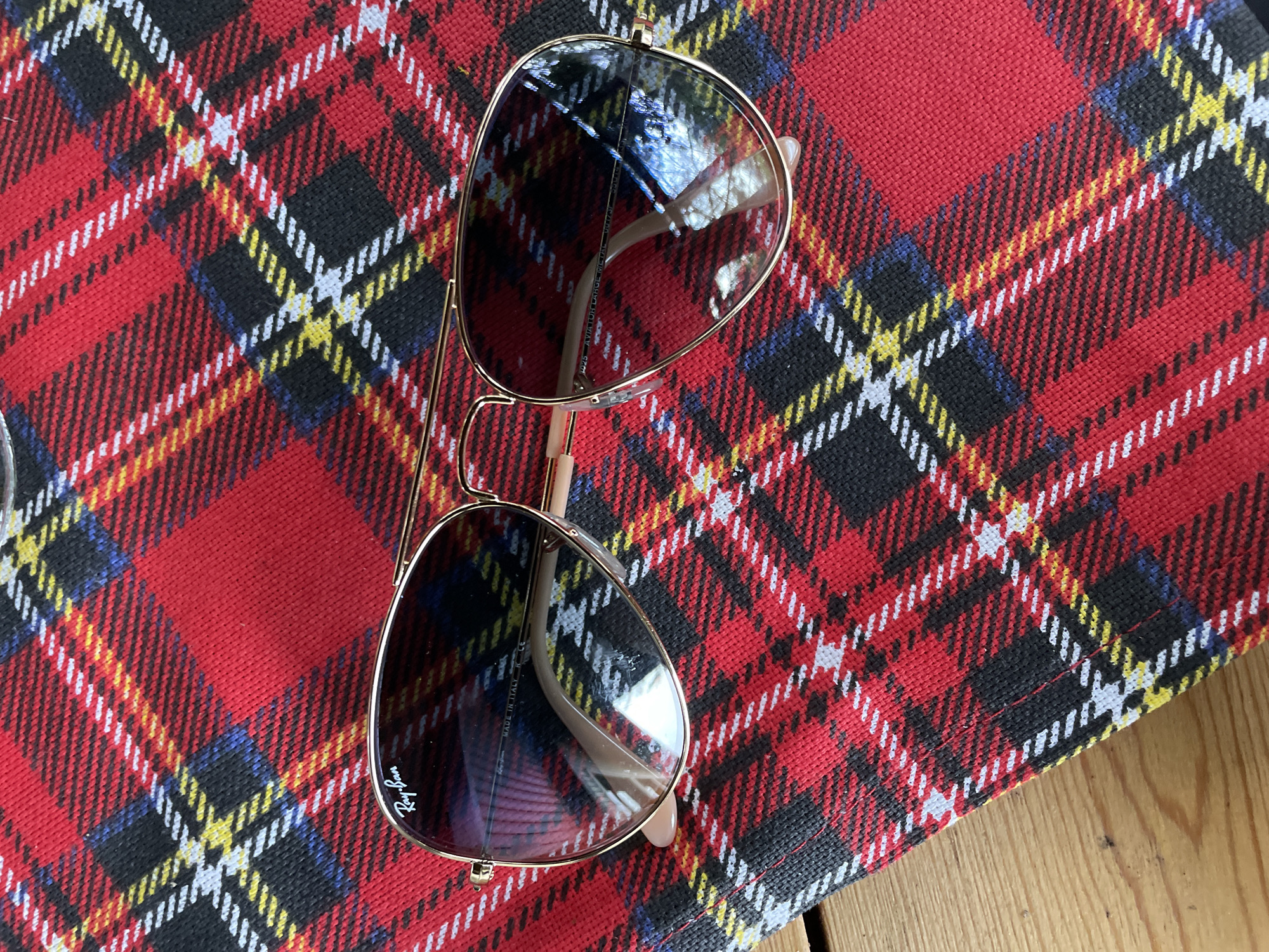 An overhead medium shot shows a pair of gold-rimmed Ray-Ban aviator sunglasses resting on a red tartan fabric. The sunglasses are positioned diagonally, with the left lens slightly higher than the right. The lenses appear to be tinted, reflecting the surrounding environment. The fabric has a repeating pattern of red, black, yellow, and white plaid lines. The lower left corner of the image reveals a glimpse of a light brown wooden surface, likely a table. The background is entirely covered by the tartan fabric. There are no people or animals present in the image. The scene is indoors, as indicated by the lighting and the presence of fabric and furniture.