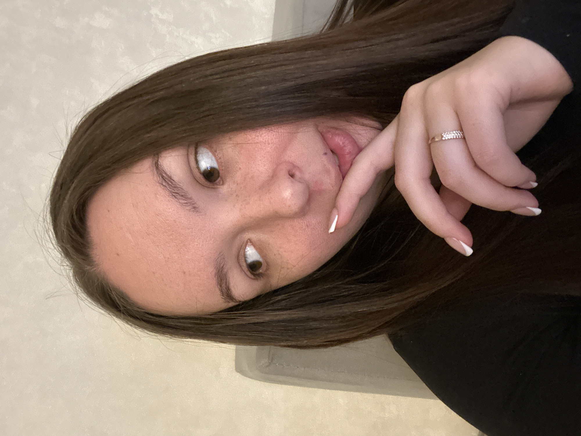 A woman with brown hair, light skin, and freckles is pictured in a close-up shot. She has a ring on her finger and is sticking her tongue out slightly. The background appears to be a light-colored wall and a gray object, likely a piece of furniture. It is not possible to determine the location as the city of Chaikovsky, Russia, from the image.