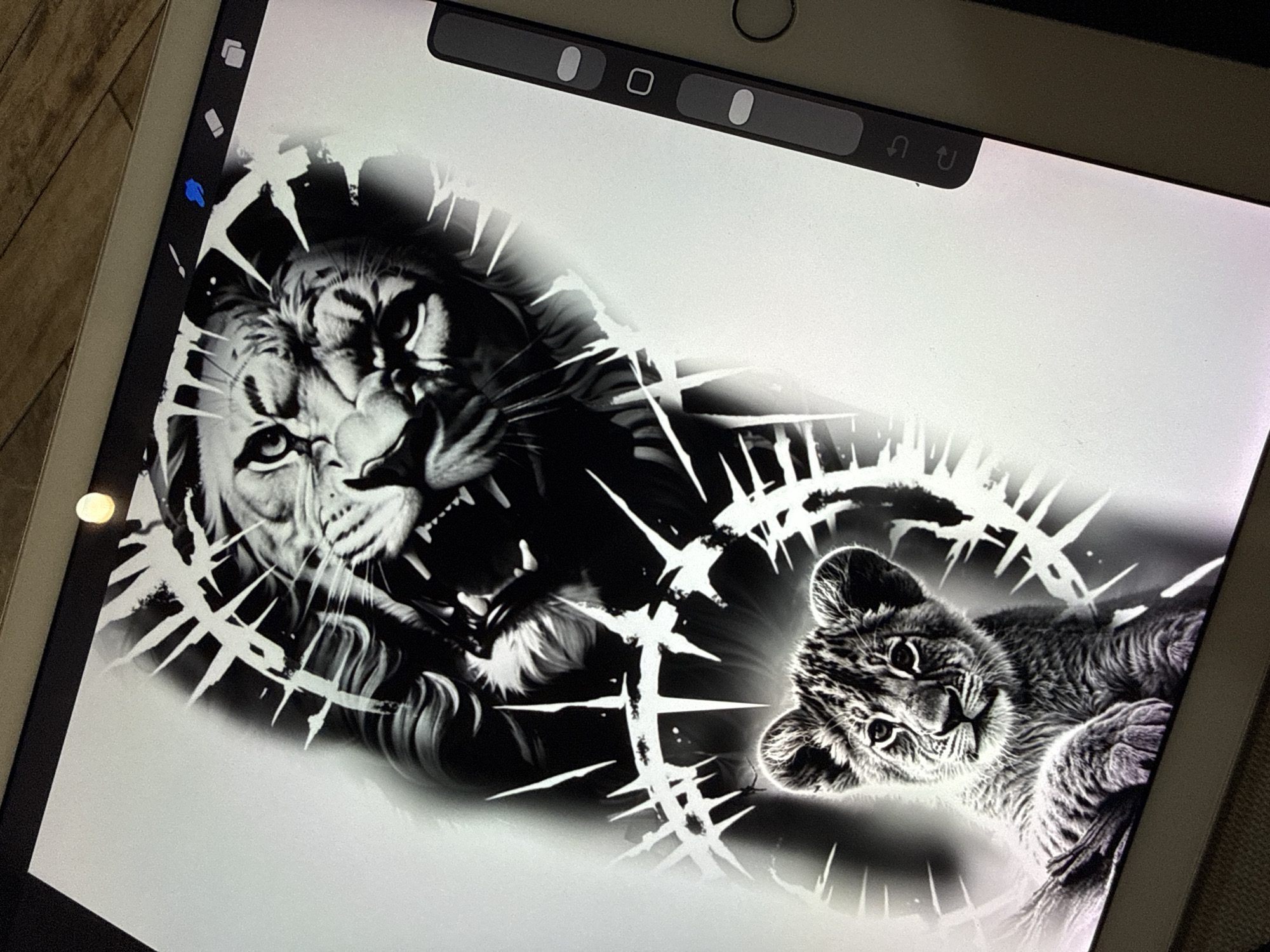 An overhead shot captures a digital artwork displayed on a tablet, likely an iPad, resting on a wooden surface in Sofia, Bulgaria. The artwork is rendered in grayscale with stark black and white contrasts, showcasing an adult lion and a lion cub amidst jagged, spiky white elements that frame them like thorns or shattered glass.
On the left, a powerful, roaring adult lion is depicted with its mouth open, fangs visible, and an intense gaze, conveying ferocity and strength. Its fur is meticulously detailed, with glowing white highlights adding depth. To the right, a much smaller, calm lion cub lies on its back or side, looking towards the viewer with an innocent expression. Its fur, like the adult's, is rendered with impressive detail and soft white highlights. The jagged white shapes serve as a dynamic backdrop, radiating outwards from the lions and creating a sense of energy or tension.
The tablet's interface is partially visible, showing typical digital art software elements. On the left side, icons for a brush, an eraser (highlighted in blue), and layers are visible. Along the top, a control bar features two sliders and a square icon, likely for brush adjustments. To the far right of this bar, undo and redo arrow icons are present. The screen's brightness and the crispness of the digital art suggest an indoor setting with adequate lighting.