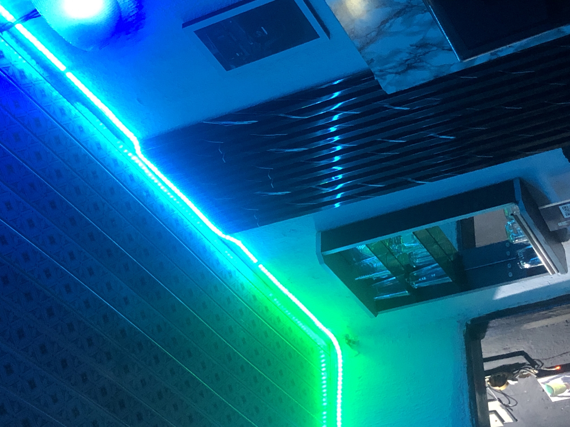 The image depicts an indoor scene illuminated by vibrant LED strip lights, casting green and blue hues. The ceiling features a textured pattern, possibly acoustical tiles, running diagonally. A wall is adorned with vertical wooden slats, creating a textured effect.
In the foreground, a dark, possibly wooden or metal structure, could be a cabinet or shelf unit, is visible. It contains several glasses. Another shelf is visible further down, also holding glasses. To the left, a doorway or opening reveals more of the interior, with some items hanging on the wall.
A framed picture or artwork is mounted on the wall to the right. The overall impression is a dimly lit, modern interior with a focus on ambient lighting. No people are present in the image. The location is unknown.