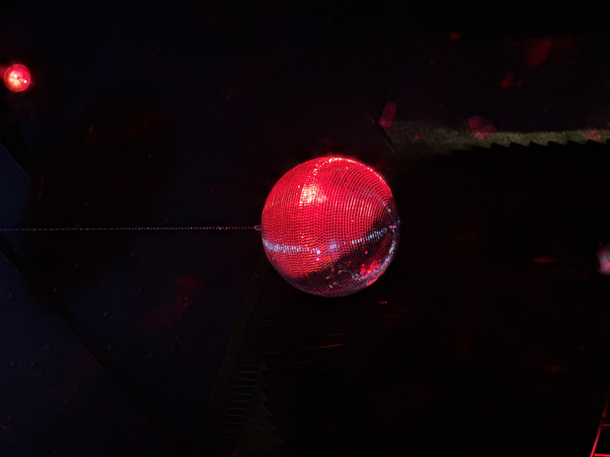 This image depicts a large, red-illuminated disco ball suspended in a dark indoor environment, likely an entertainment venue in Sofia, Bulgaria. The central disco ball is covered with small, mirrored tiles that brightly reflect red light, creating a shimmering effect. A distinct white highlight is visible on its surface, indicating the primary light source.
To the upper left of the main subject, a smaller, round red light source or orb is visible, glowing softly against the dark background. A thin, metallic chain extends horizontally from the disco ball towards the left edge of the frame, suggesting how it is suspended.
The background is largely shrouded in darkness, with faint, indistinct shapes and structures visible. These dark elements appear to be architectural features or stage rigging, subtly touched by red ambient light. In the upper right corner, a series of jagged, sawtooth-like patterns are discernible, possibly hinting at stairs, a railing, or part of the ceiling structure. The scene evokes the atmosphere of a nightclub, party, or concert, characterized by its dramatic lighting and celebratory object. There is no visible text in the image.