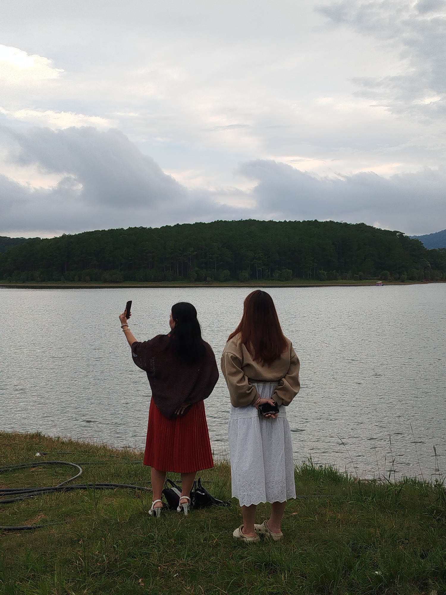 Two women stand on a grassy bank overlooking a large lake. The woman on the left, facing away from the camera, is wearing a dark brown shawl over a red pleated skirt and white sandals. She is holding up a smartphone in her right hand, appearing to take a photo or selfie. The woman on the right, also facing away, is wearing a tan sweatshirt over a white lace-trimmed dress and light-colored slip-on shoes. She has her hands clasped behind her back. A black bag rests on the grass between them. In the background, a dense forest covers a hillside that slopes down to the water's edge. The sky is overcast with heavy clouds. The overall scene suggests a peaceful, natural setting, possibly during a trip or outing. The time of day appears to be late afternoon or early evening, given the diffused light. The location context is Da Lat, Vietnam. There is no visible text in the image.