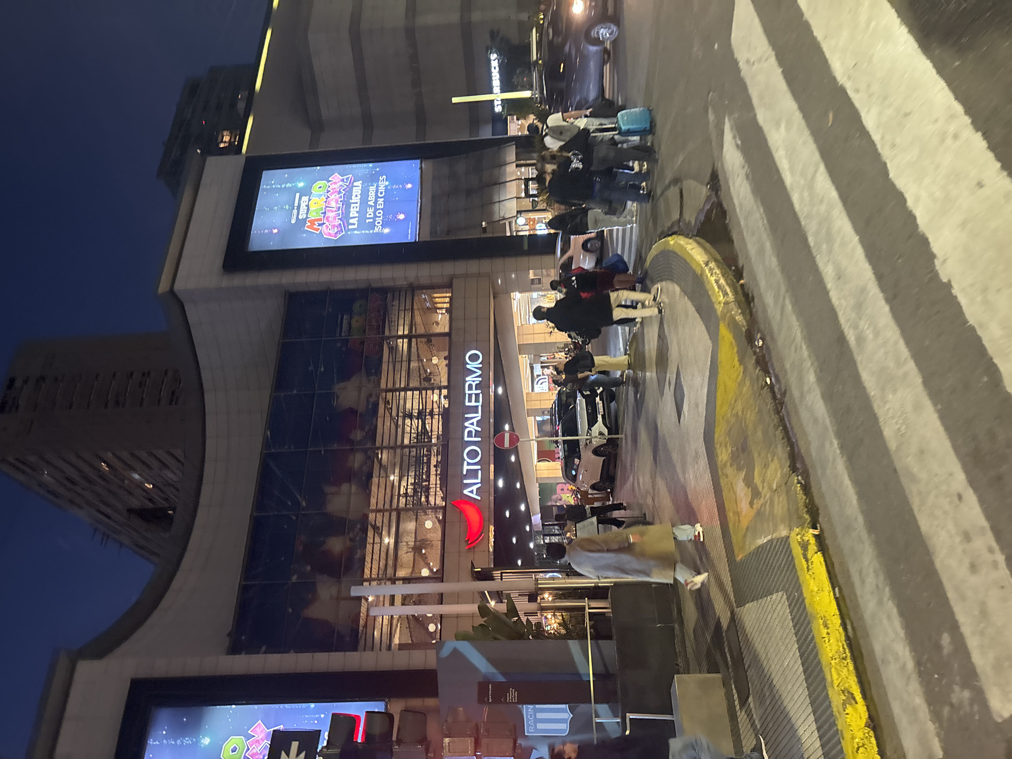 An evening scene depicts the exterior of a large, modern shopping mall, identified by the prominent "ALTO PALERMO" sign, in an urban setting. The sky is dark blue, indicating dusk or night, with the building and surrounding area illuminated by artificial lights and large digital screens.
Numerous people, including pedestrians, are visible on the sidewalk and crossing the street on a marked crosswalk. Some individuals appear to be waiting near the mall's entrance, while others carry luggage, suggesting arrival or departure from a trip. Cars are also present on the street.
Notable features of the building include large glass windows reflecting the interior lights, and two prominent digital billboards. One billboard clearly displays an advertisement for "SUPER MARIO BROS. LA PELÍCULA," with text below reading "1 DE ABRIL SOLO EN CINES." Another sign for "STARBUCKS" is visible higher up on an adjacent structure. A partially visible sign at the bottom of the image shows "RACIN." The street features a zebra-striped crosswalk and a yellow-painted curb. The overall atmosphere is busy but orderly, characteristic of a commercial area at night.