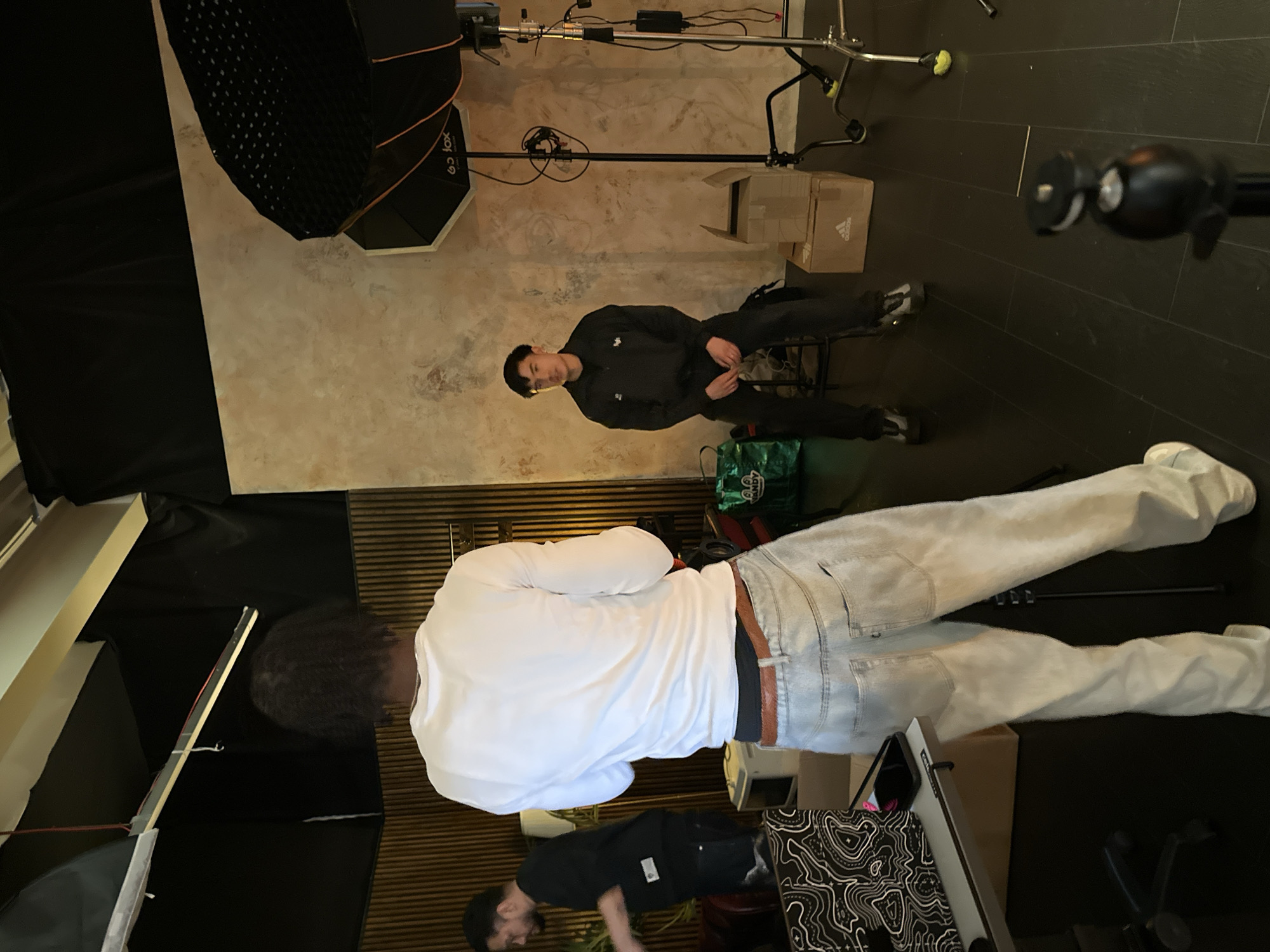 The image shows an indoor scene with three people and photographic equipment. One person is lying down, facing away from the camera, wearing a white long-sleeved shirt and light-wash jeans. Another person is seated, facing the camera, wearing a dark tracksuit. A third person is bent over in the lower portion of the frame wearing a dark shirt.
Lighting equipment, including a softbox on a stand, is positioned near the top of the frame, suggesting a photography or video shoot setting. A cardboard box with Adidas branding is visible near the top right. A patterned bag or cloth lies between the seated person and the person lying down.
The setting appears to be a studio or indoor location. There are dark wooden slats behind the person in white, a white wall is behind the person in the dark tracksuit, and dark tile floor is at the top right.
Given the Adidas branding and the presence of studio equipment, this could be a photography session for promotional materials. The specific location in London, United Kingdom, is not discernible from the visual information provided.
