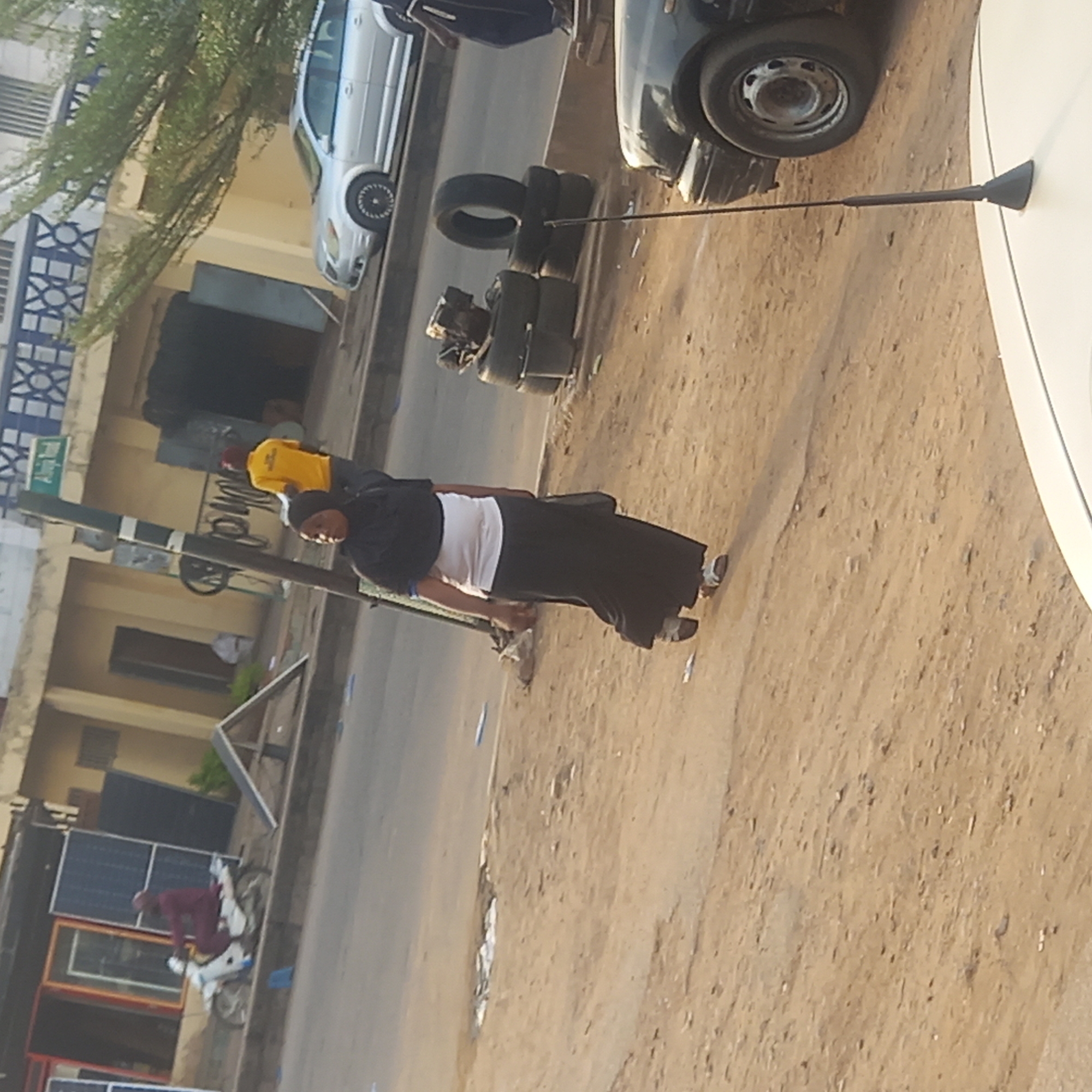 A street-level view, possibly Dadi Riba, Nigeria, depicts a road with various objects and people. A woman in a black head covering and a white shirt with a black skirt is walking on the road's edge. A man in a yellow shirt is walking nearby. Several stacked tires are present on the road. Two cars, one silver and one black, are parked along the roadside. In the background, there are buildings, including one with solar panels displayed in front. A man sits on a motorcycle nearby. The road surface is dirt.