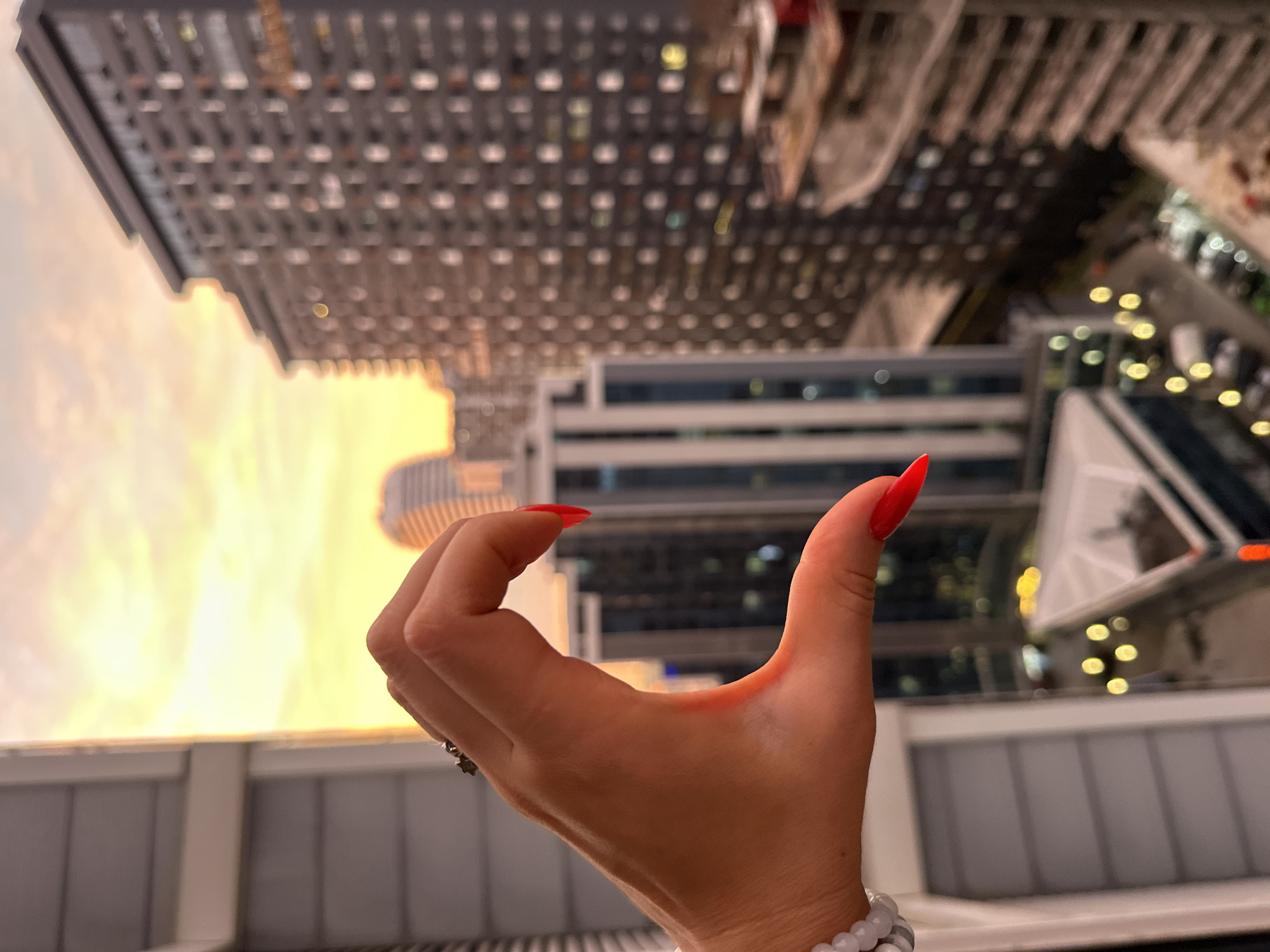 The image shows a person's hand with bright red, almond-shaped nails, forming a "C" shape. The hand is positioned in the foreground, against a backdrop of blurred city buildings and a bright, warm sky, suggesting it is either sunrise or sunset. The buildings appear to be modern high-rises, with many windows reflecting the light. The overall scene is slightly abstract due to the blur and the inverted perspective, which makes the buildings seem to hang from the sky. The lighting and the warm glow of the sky evoke a sense of calm or perhaps contemplation. The location is identified as Batumi, Georgia, known for its contemporary architecture and coastal setting, which aligns with the visual elements of the image. The image does not contain any visible text.