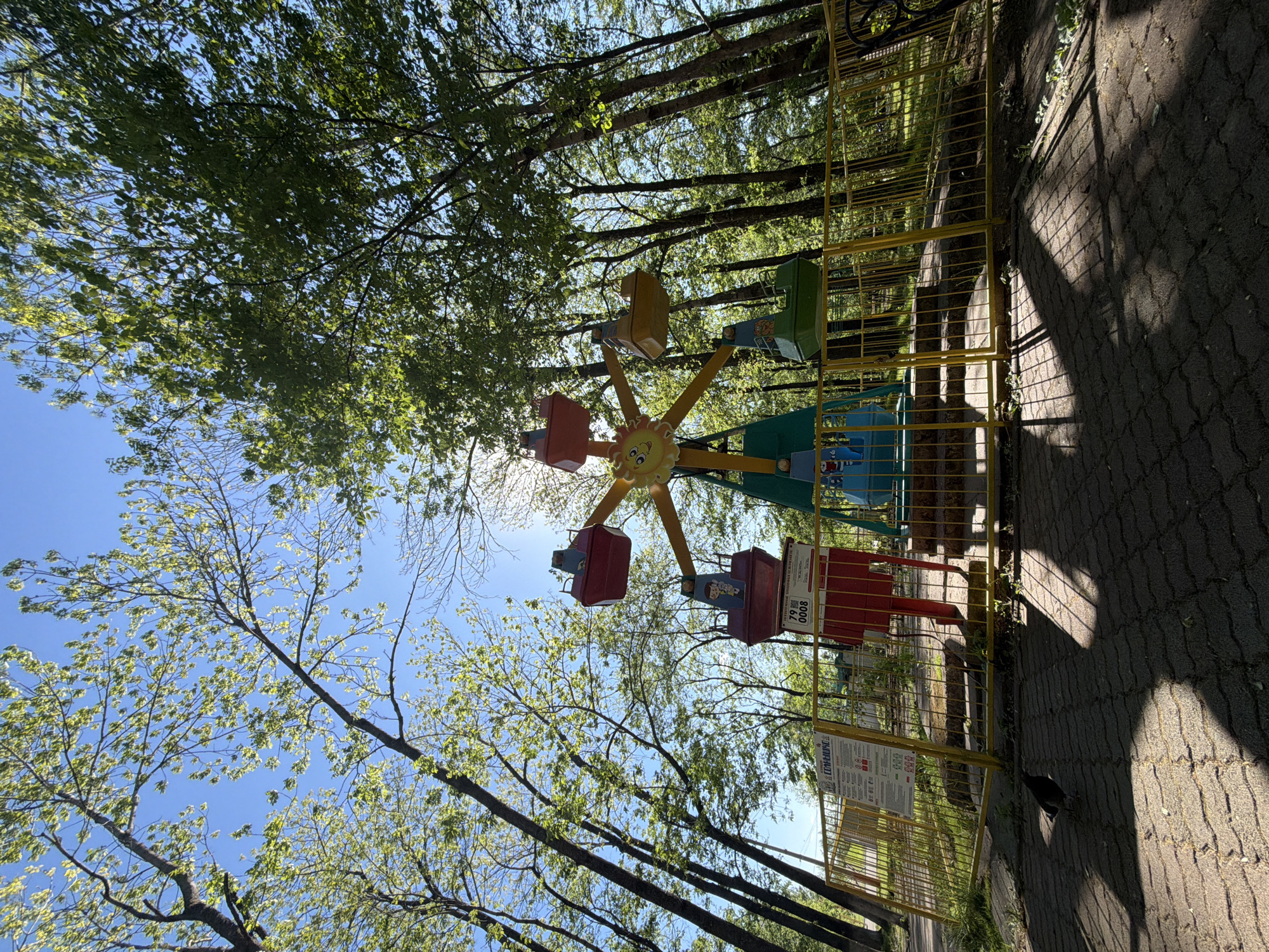 The image shows a children's amusement park ride in Birobidzhan, Russia. The ride is a small Ferris wheel with brightly colored cars attached to a central yellow sun-shaped hub. The ride is surrounded by a yellow metal fence. Behind the fence, there are trees with green leaves against a blue sky. A paved walkway is visible in the foreground, with shadows cast by the fence and trees. There is also a sign with writing on it near the fence.