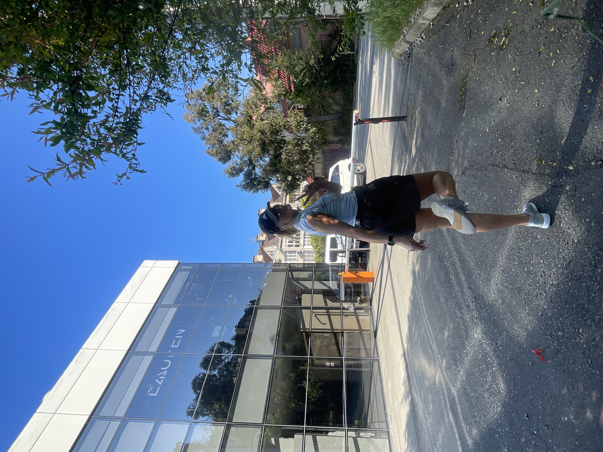This media file, originally rotated 90 degrees clockwise, captures a bright, sunny outdoor scene in what appears to be an urban or suburban setting. The time of day is clearly mid-day or early afternoon, evidenced by the clear blue sky and strong shadows cast by trees and buildings.
The central subject is a person, likely a man, standing casually in the lower-right portion of the frame. He is wearing a light-colored t-shirt, dark shorts, a dark baseball cap worn backward, and light-colored sneakers. A watch is visible on his left wrist. His left hand is raised, making a peace sign gesture towards the camera, suggesting a relaxed or positive mood.
To the left and behind the person is a prominent modern building with a multi-story glass facade, which reflects the blue sky and nearby trees. The word "LAUTEN" is visible in white letters, reflected in reverse on one of the glass panels. The foreground consists of an asphalt road or pavement with scattered leaves and a small red object on the ground.
In the mid-ground, a white car is partially visible near a black and white barrier arm. Further into the background, older, traditional-style houses with reddish-brown roofs are nestled among numerous green trees and dense foliage, some of which overhang the scene from the upper left, providing dappled shade. The combination of modern architecture, traditional housing, and lush greenery suggests a diverse urban landscape, consistent with the location context of Sochi, Russia.