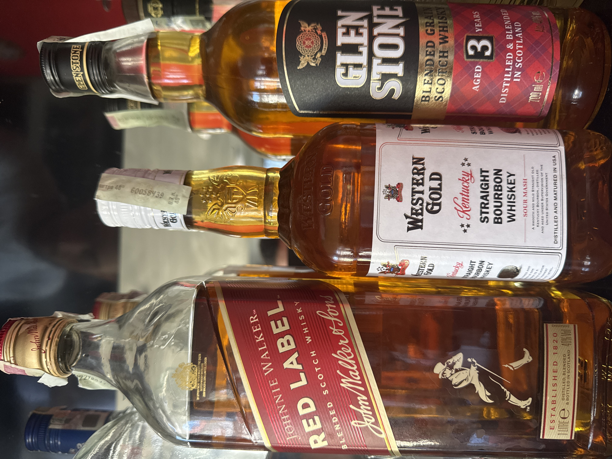 The image shows three bottles of liquor arranged in a stack on a reflective surface. The bottom bottle is Johnnie Walker Red Label Blended Scotch Whisky. The middle bottle is Western Gold Kentucky Straight Bourbon Whiskey. The top bottle is Glen Stone Blended Grain Scotch Whisky, aged 3 years. The location in Dermantsi, Bulgaria is not provided and not apparent.