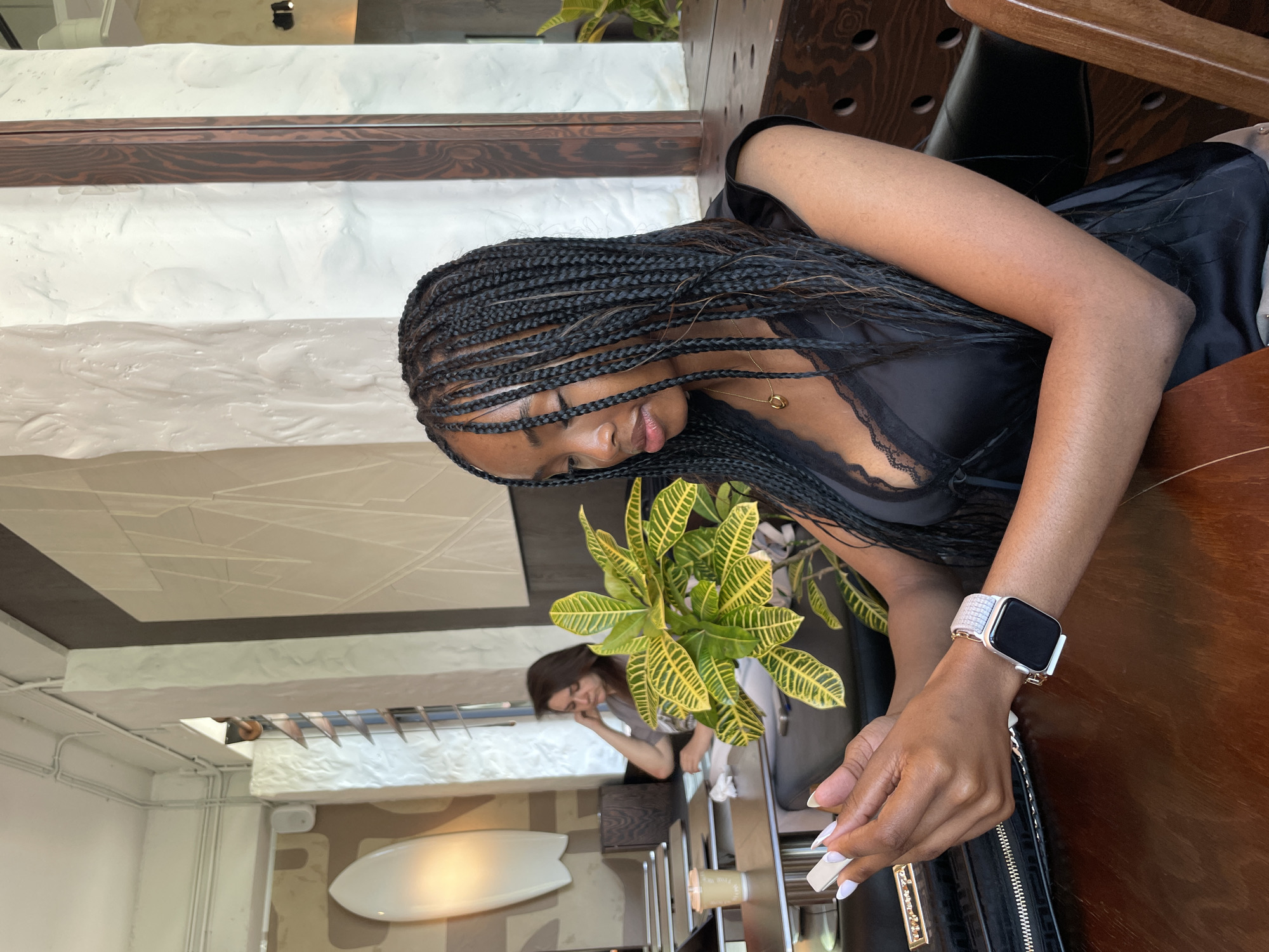 A Black woman with braided hair, wearing a black top with lace trim, is seated at a wooden table in a cafe and looking at her phone. She has a gold necklace and a smartwatch on her left wrist. A black clutch bag is on the table in front of her. In the background, another woman with brown hair is seated at a table, looking down and resting her head on her hand. There is a potted plant with green and yellow leaves on the table between the two women. The cafe has textured white walls, a wooden beam, and what appears to be a surfboard-shaped object on the wall. The overall setting suggests a modern cafe environment. The location of Kazan, Russia is not discernible from the image.