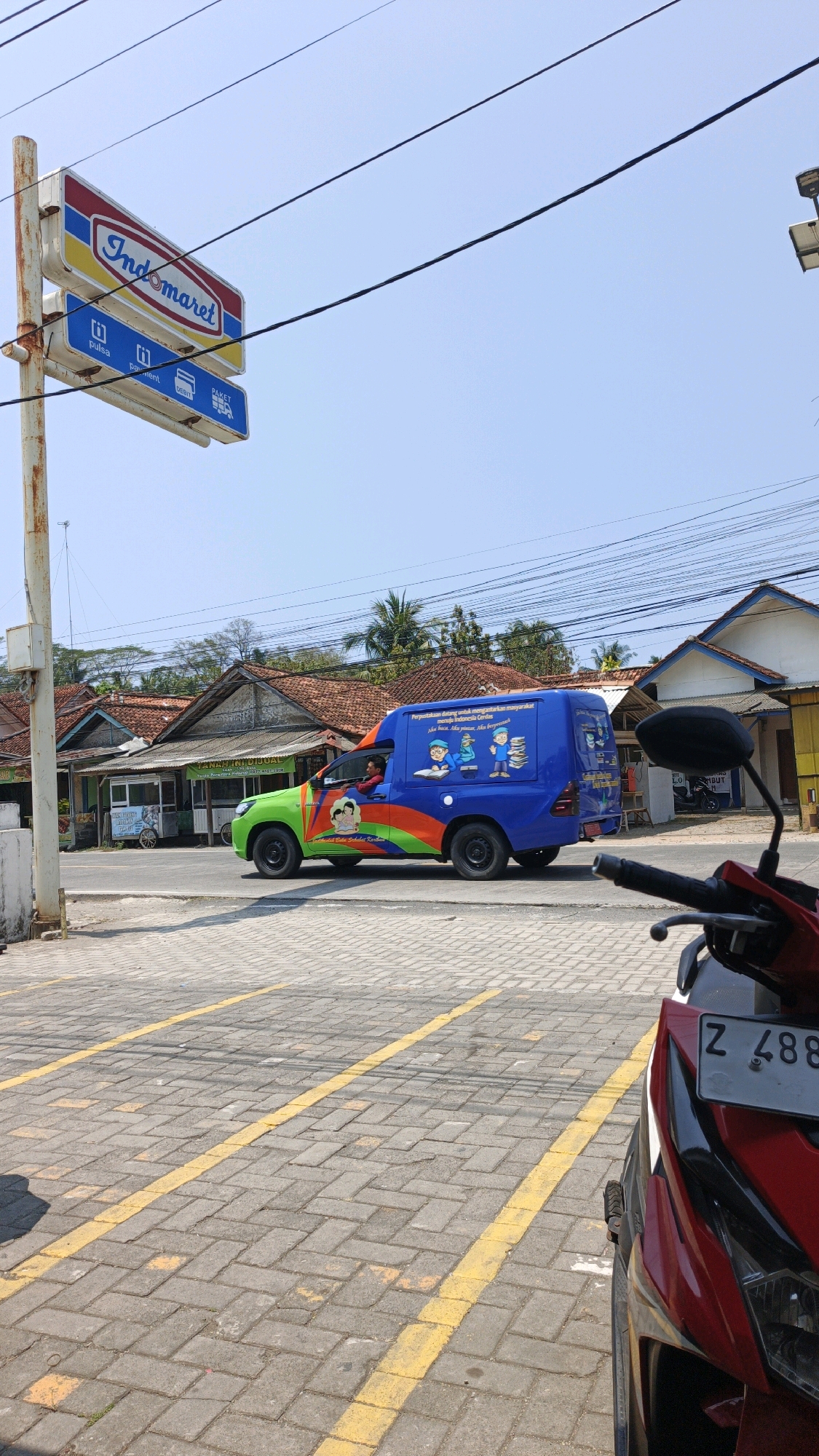 An outdoor, eye-level, medium shot captures a brightly colored blue and green van with orange and red accents driving on a paved road in Cibenda, Indonesia. The van, decorated with illustrations of children reading and a book, appears to be a mobile library. A driver wearing a red shirt is visible inside the van. To the left of the road, an Indomaret sign is mounted on a pole, indicating a convenience store. Behind the van, several traditional Indonesian houses with tiled roofs are visible, along with palm trees and utility poles with power lines. In the foreground, a red motorcycle is partially visible, with its license plate reading "Z 488". The ground is paved with interlocking bricks and marked with yellow lines, suggesting a parking area or a designated road section. The sky is clear and blue.