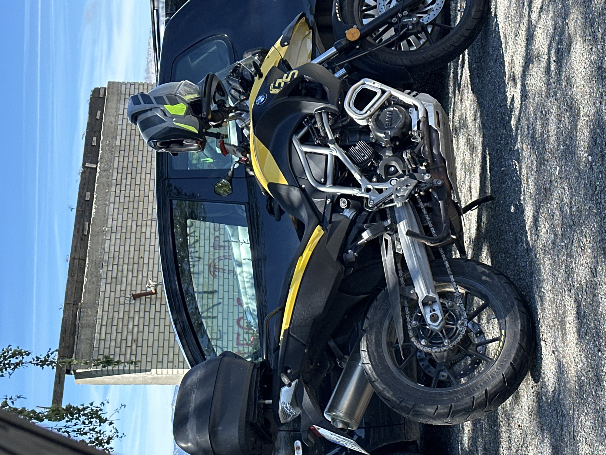 A yellow and black BMW F 750 GS motorcycle is parked on a gravel surface in front of a dark-colored car. The motorcycle has a black top case, a silver exhaust pipe, and a helmet with gray and neon yellow accents resting on its handlebars. In the background, there is a brick wall, a wooden beam, and a clear blue sky. The scene is located in Jabuka, Montenegro, under bright daylight.