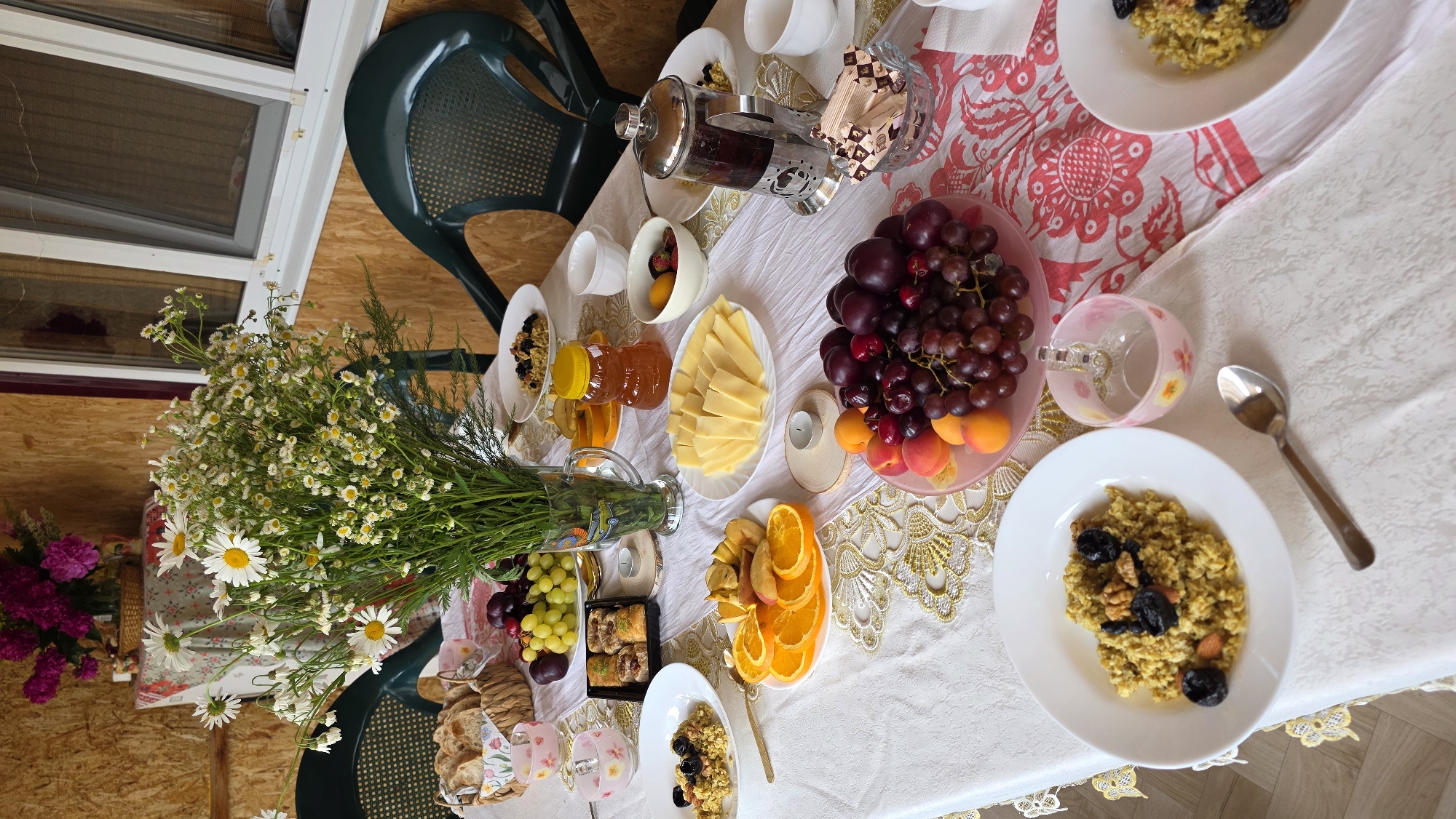 The image depicts an outdoor table setting, likely on a porch or veranda, with a spread of breakfast items. The table is covered with a white tablecloth adorned with a gold lace trim and a pink floral pattern on a portion of it.
In the center of the table, a large bouquet of white daisies and smaller white flowers in a glass vase stands prominently. To the left of the flowers, a woven basket contains sliced bread. Several small pink glass cups with floral patterns are also present.
The food items arranged on the table include plates of porridge topped with nuts and dried fruit, sliced oranges and apples, a bowl of grapes, plums, and apricots, a tray of baklava, sliced cheese, a jar of honey, a bowl of berries and fruit, and a French press filled with dark liquid, presumably coffee or tea.
Two green plastic chairs are visible on either side of the table, suggesting outdoor seating. The background shows a wooden wall and a window with a screen. The overall scene suggests a pleasant breakfast or brunch enjoyed outdoors, possibly in a rural or suburban setting given the natural elements and simple furnishings. The location is specified as Kupoyarovo, Russia. There are no people visible in the image.