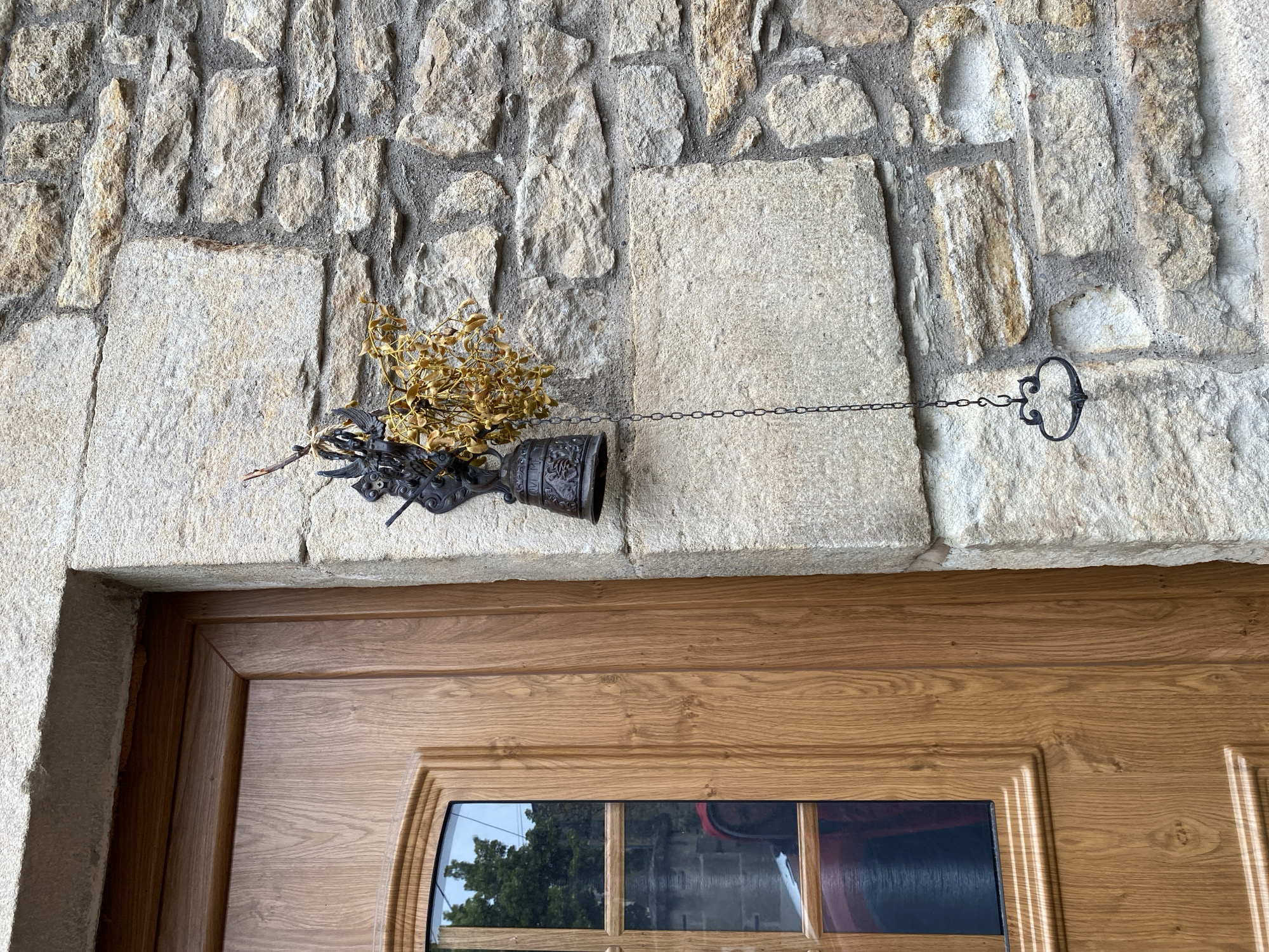 The image displays a rustic architectural detail of a building entrance in Billom, France, focusing on a stone wall above a wooden door. The upper portion of the frame is dominated by a wall constructed from irregular, rough-hewn stone blocks in shades of light grey and beige, interspersed with dark mortar.
Mounted on this stone wall, centrally above the door, is a decorative object. It features a dark, ornate metal base resembling an inverted bell or small chalice, from which a bundle of dried, golden-brown foliage or branches protrudes upwards and outwards. Extending horizontally to the right from this object is a slender, dark metal chain. This chain terminates at a smaller, intricately curled, C-shaped metal hook or bracket, which is securely attached to the stone wall.
Below the stone wall, the top section of a sturdy wooden door is visible. The door is crafted from medium-brown wood with a distinct vertical grain pattern, and it features carved, recessed panels. Towards the bottom of the visible door area, there are rectangular glass inserts that reflect the surrounding environment, showing glimpses of green trees and hints of other distant objects, indicating a daytime setting. No people are present, and no text is visible.
