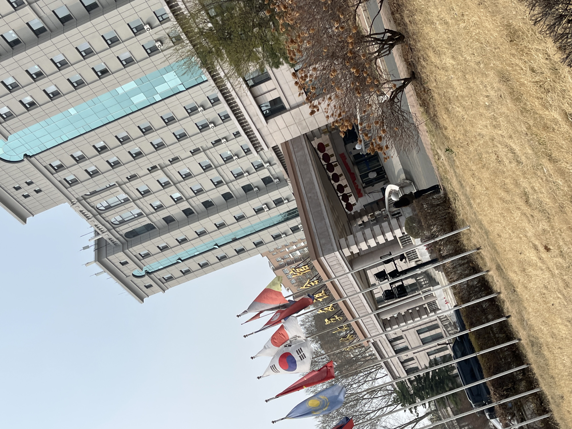 The image depicts a building with numerous windows and what appears to be an architectural glass feature. Several flags, including those of South Korea, Japan, Kazakhstan and China, are displayed in front of the building. The surrounding environment includes trees, a grassy area, and the scene is likely located in Changchun City, China, given the flags displayed. There are people located in front of the building.