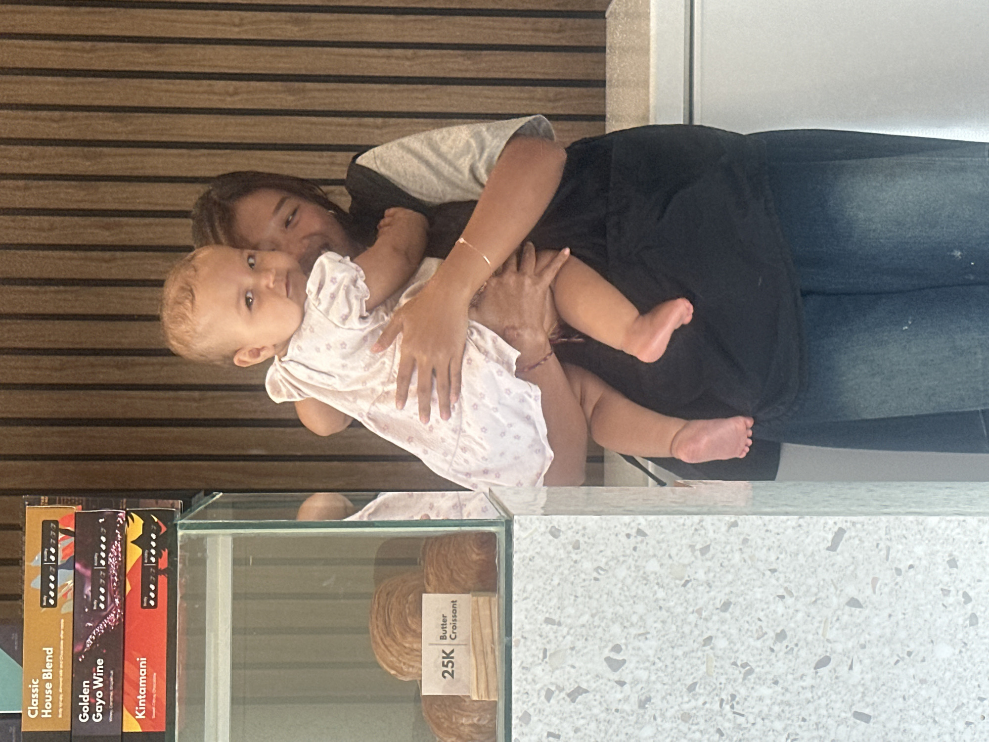 The image depicts a woman holding a baby in what appears to be a cafe or bakery in North Kuta, Indonesia. The woman is wearing a gray top, black shorts/skirt, and jeans. The baby is wearing a white dress with small floral prints. The woman is holding the baby close to her body, facing the camera. A glass display case is visible in the foreground, containing baked goods labeled "Butter Croissant" with a price of 25K. Next to the display are coffee product boxes. The background has a wooden panel design. The overall setting suggests a casual, public space.