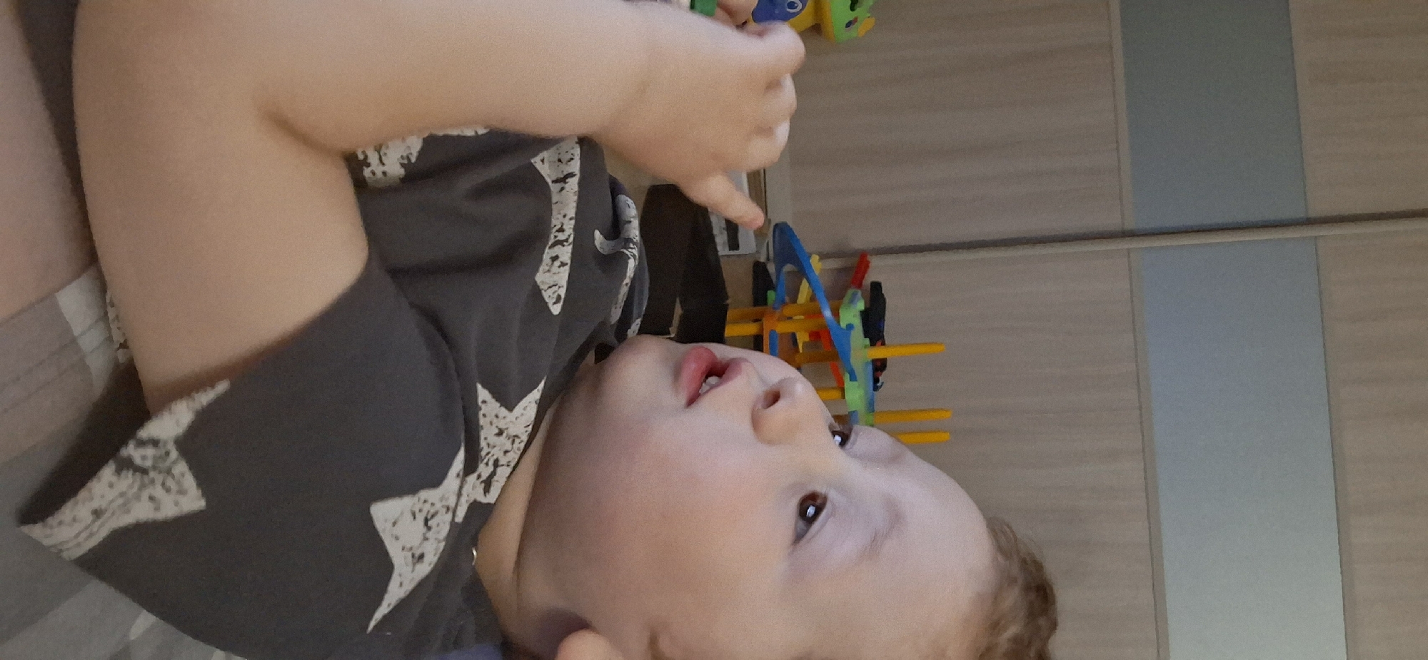 A young child is lying down wearing a gray shirt with white star patterns. The child's face is visible, with light skin, brown eyes, and open lips. The child's arm is raised, and their fingers are pointing. In the background, there are children's toys including a green and yellow toy, and a colorful construction toy with blue, green, red, and yellow elements. The background also includes a wall with light brown and gray panels. The location is Chaikovsky, Russia.