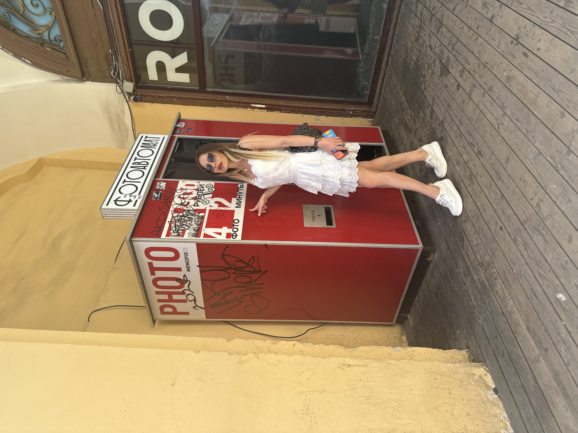 A young woman in a white lace dress and white sneakers stands in front of a red photo booth. She is wearing sunglasses and carrying a black backpack. The photo booth has "PHOTO" written on it in large red letters and "ФОТОАВТОМАТ" in white letters on a black sign above. There is graffiti on the side of the photo booth. To the right of the photo booth is a shop window with "RO" in large white letters. The scene appears to be in an outdoor urban setting, possibly in Saint Petersburg, Russia, given the Cyrillic text on the photo booth. The ground is made of wooden planks. The background wall is painted yellow.