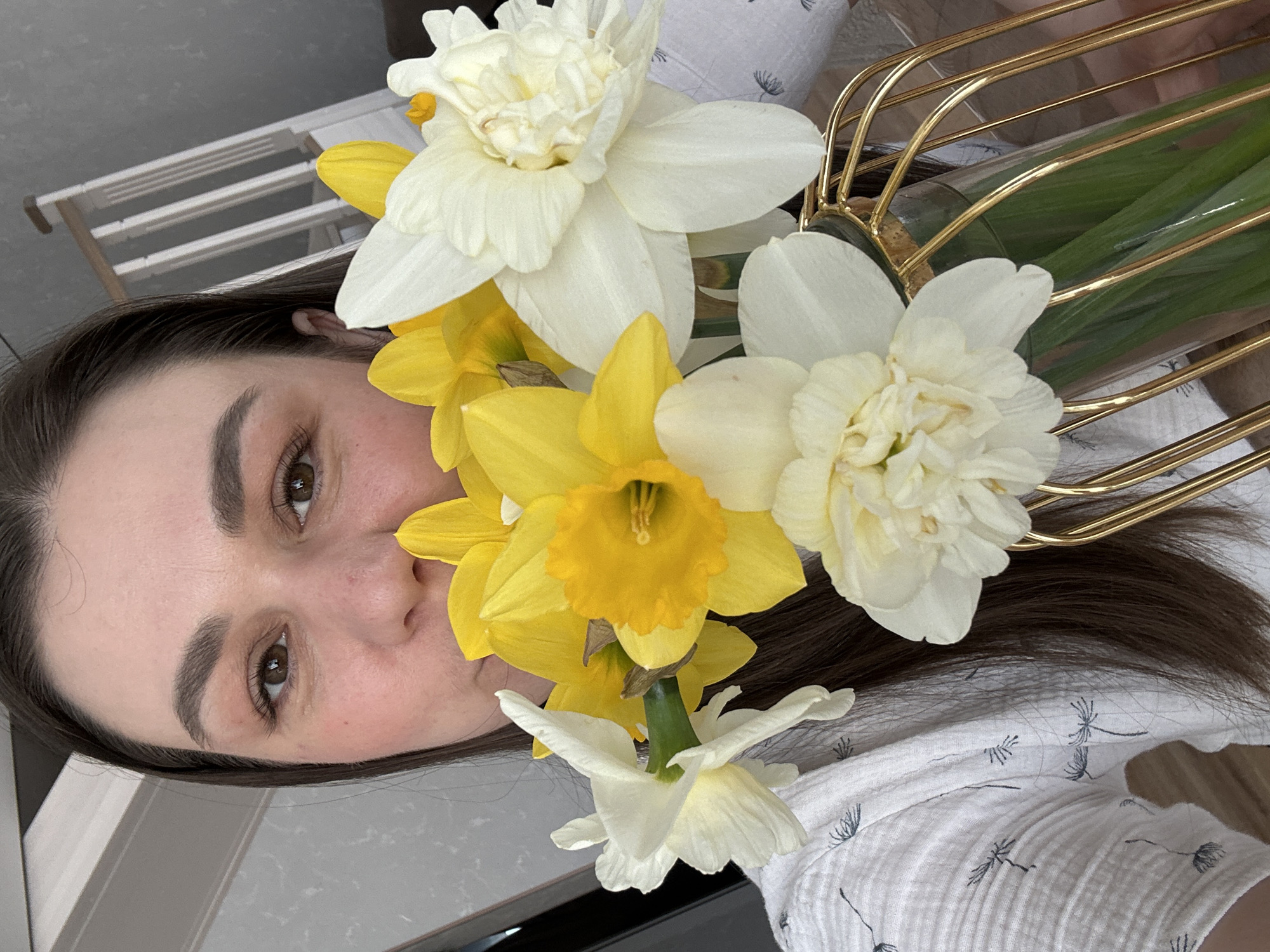 A woman is holding a vase of daffodils in front of her face. The daffodils are a mix of yellow and white. The vase is gold and has a clear glass bottom. The woman is wearing a white shirt with a dandelion print. The background is a gray countertop and a white wall with a window. The location is likely Chaikovsky, Russia, given the user's prompt.