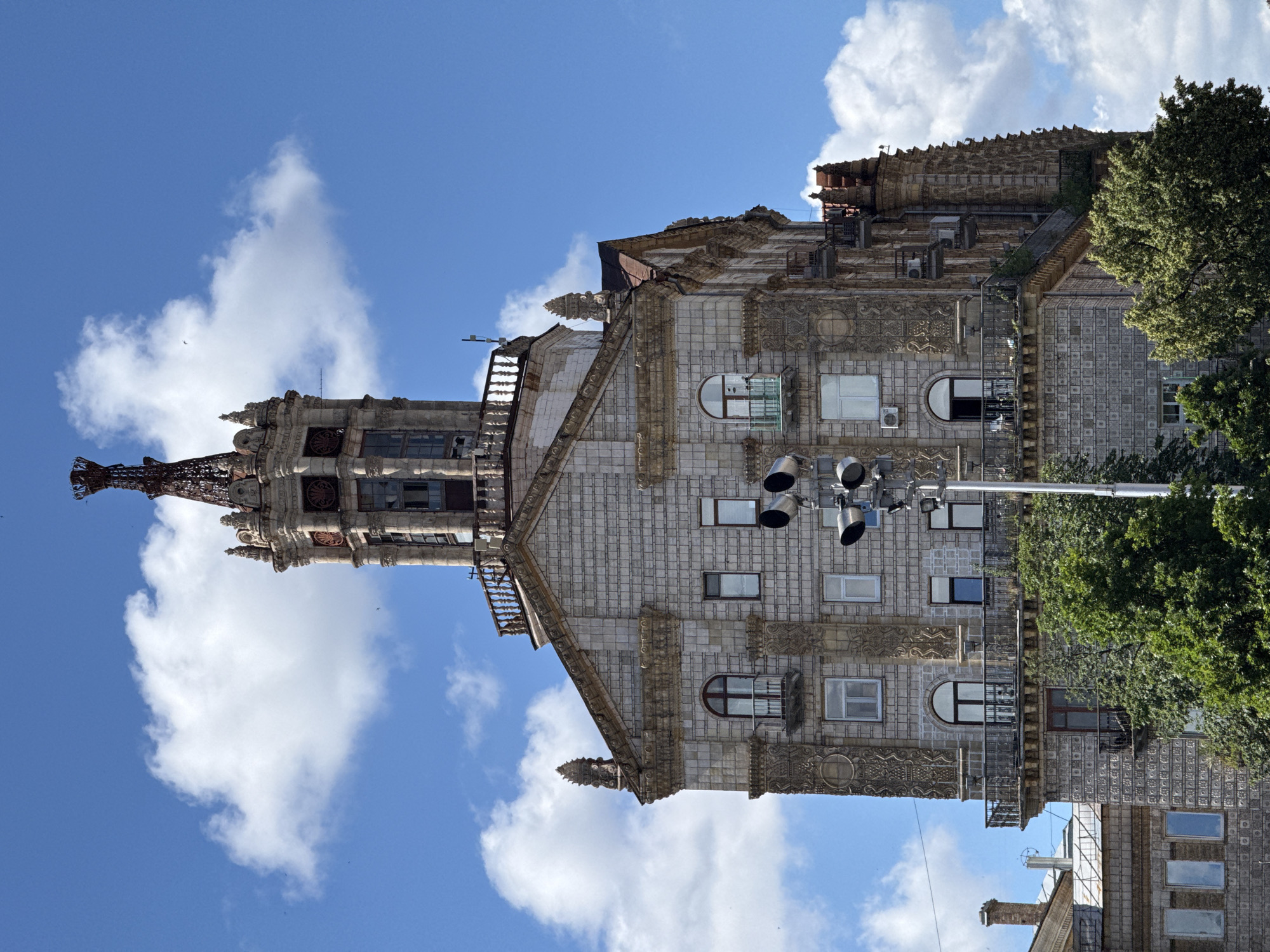 The image shows a tall, ornate building with a detailed facade and a spired tower, set against a bright blue sky with scattered white clouds. The building's exterior is covered in textured square tiles, with decorative elements around the windows and balconies. Arched windows with dark frames and small balconies with metal railings are visible on multiple levels. At the very top, a complex, wrought-iron spire crowns the structure. In the foreground, lush green trees partially obscure the lower floors of the building. A traffic light pole with multiple lights stands to the right of the building, in front of its facade.
This building is likely the House of Actors, located on Khreshchatyk Street in Kyiv, Ukraine.