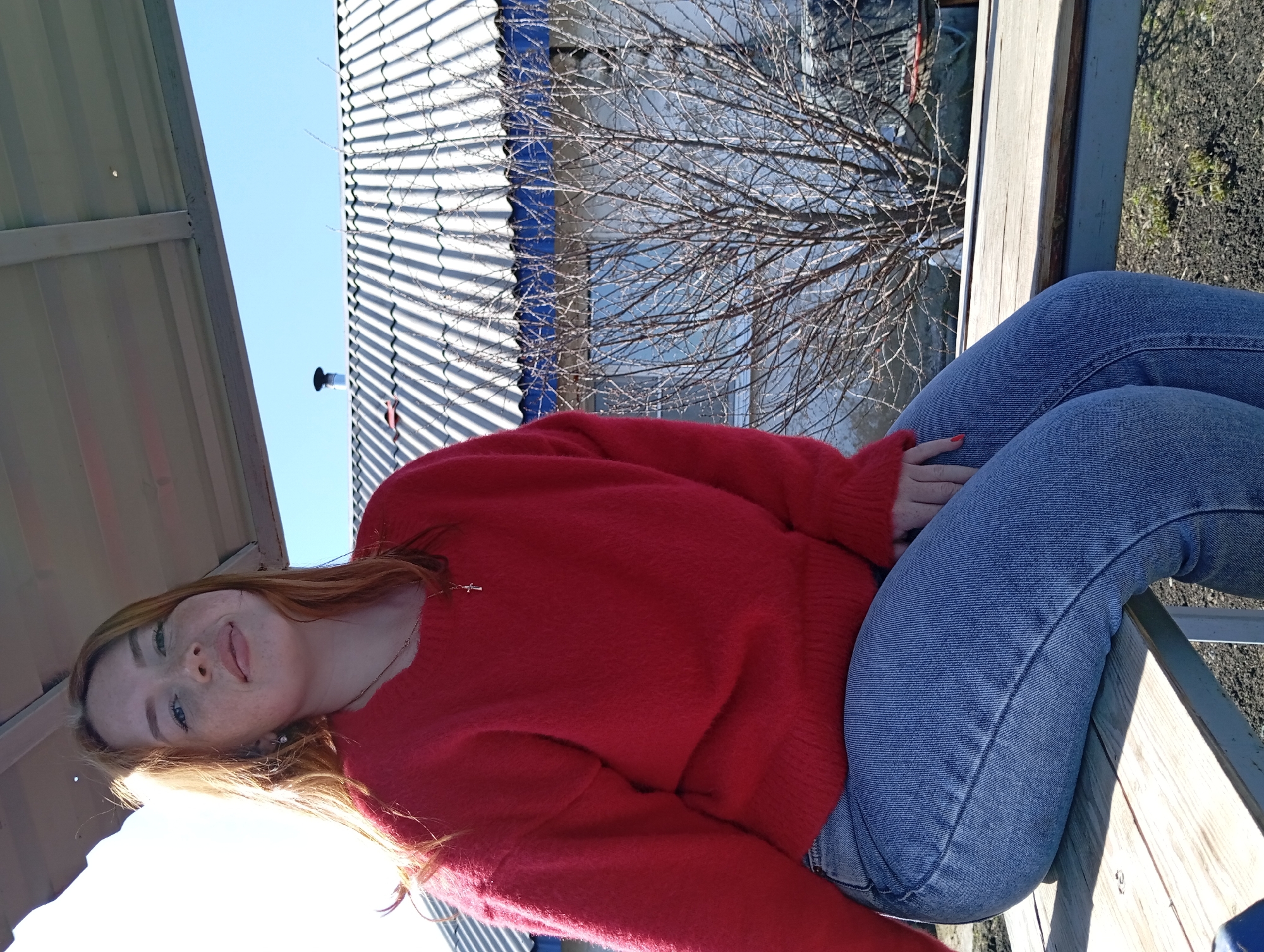 A woman is sitting on a bench in an outdoor location in Yurgamysh, Russia. She is wearing a red sweater and blue jeans. The background includes a building with a white and blue roof and a bare tree.