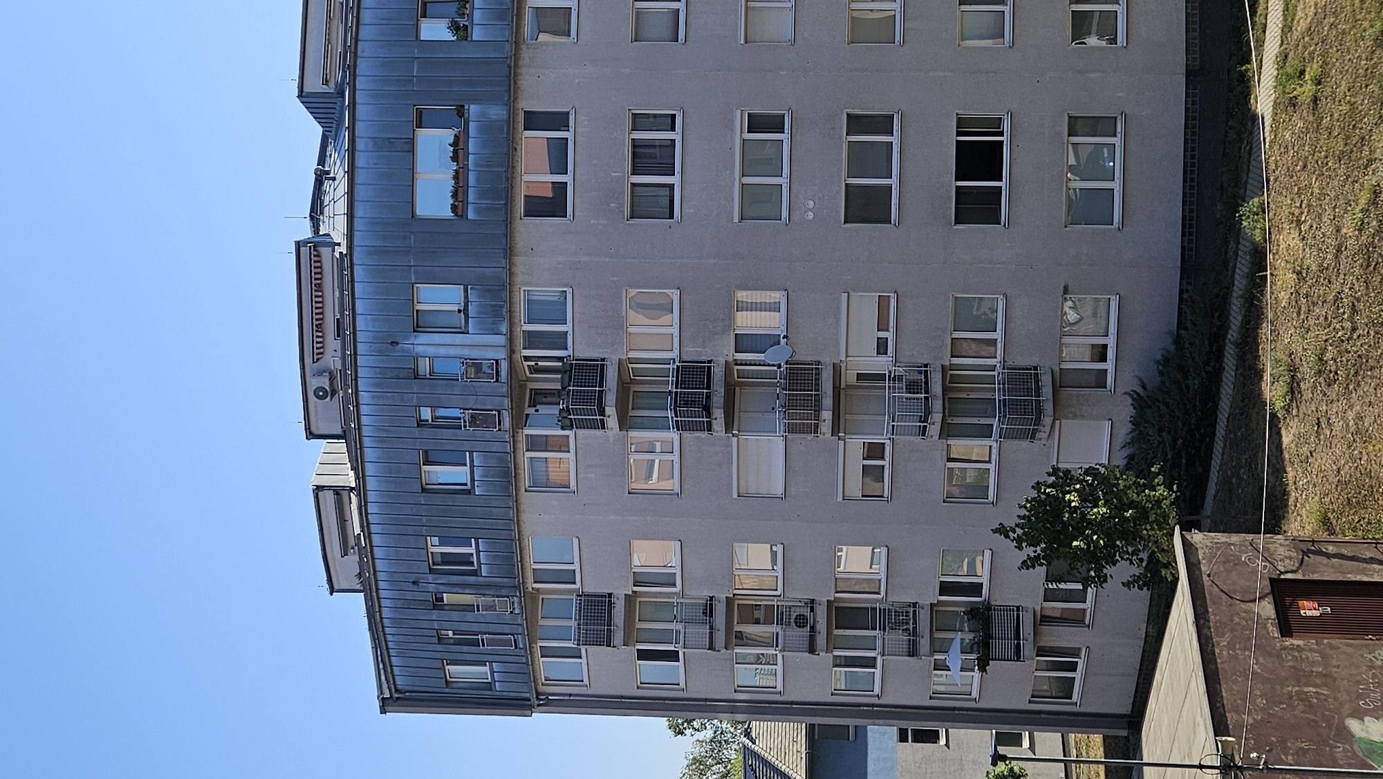 The image shows a section of a grey, multi-story apartment building with a distinctive curved facade and a metallic roof structure. The building has numerous windows and balconies. The sky is clear and blue. In the foreground, there is a low, dark-colored building with graffiti on its side, and a patch of dry, grassy ground with a walkway. To the left of the apartment building, a smaller blue building is partially visible, and trees and greenery are present. The scene is set in Bratislava, Slovakia.