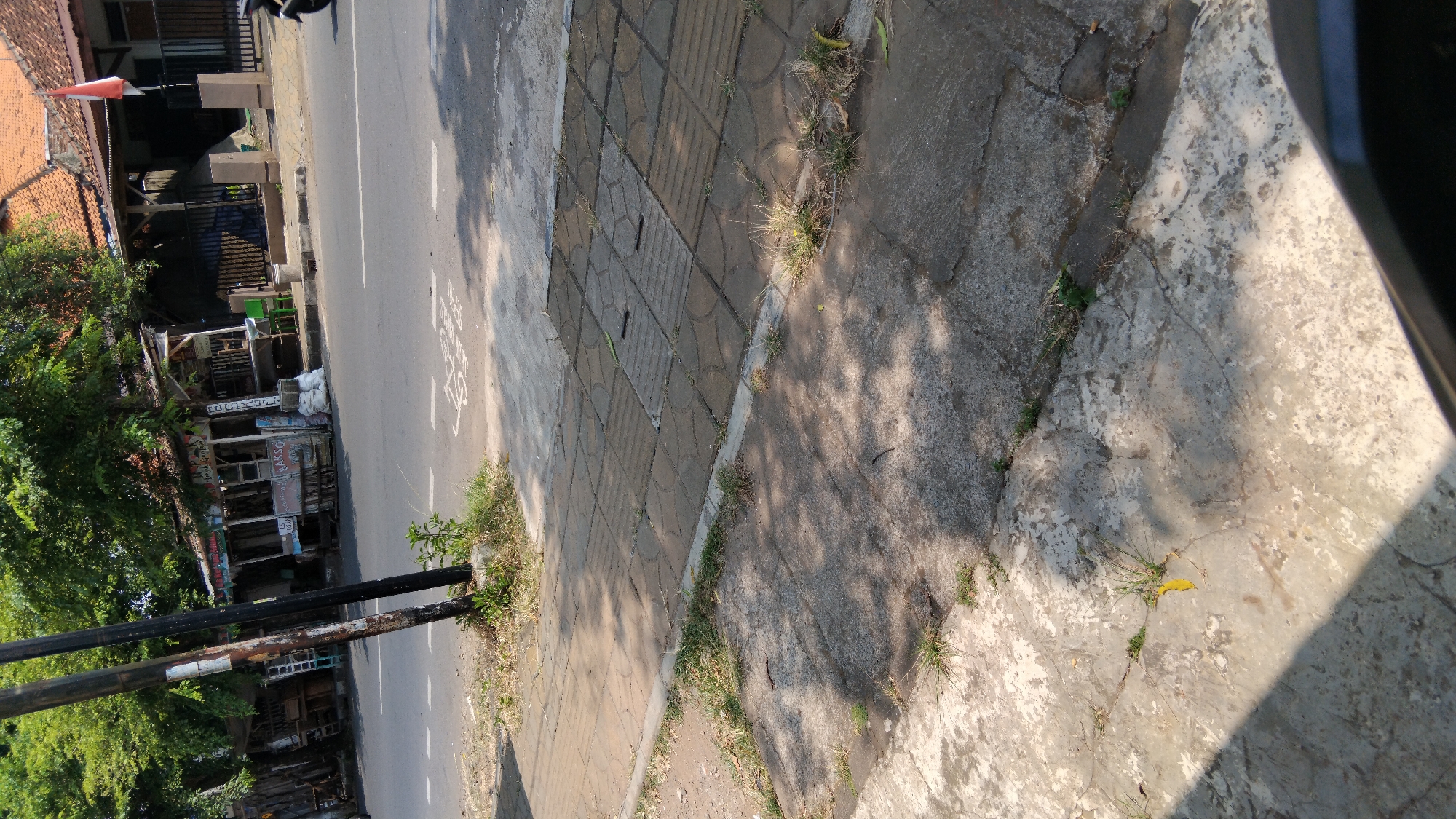 This image depicts a sidewalk next to a road in Proyonangg, Indonesia. The sidewalk is made of concrete paving stones and some sections are worn and cracked. Weeds are growing through the cracks in the sidewalk. To the left of the sidewalk is a dark pole, possibly for a street lamp. Beyond the sidewalk is a paved road with white lane markings. On the right side of the road, there are small food stalls and buildings with red tile roofs. A flag, appearing to be the Indonesian flag, is visible on a pole near one of the buildings. The scene is illuminated by sunlight, casting shadows from trees onto the sidewalk and road.