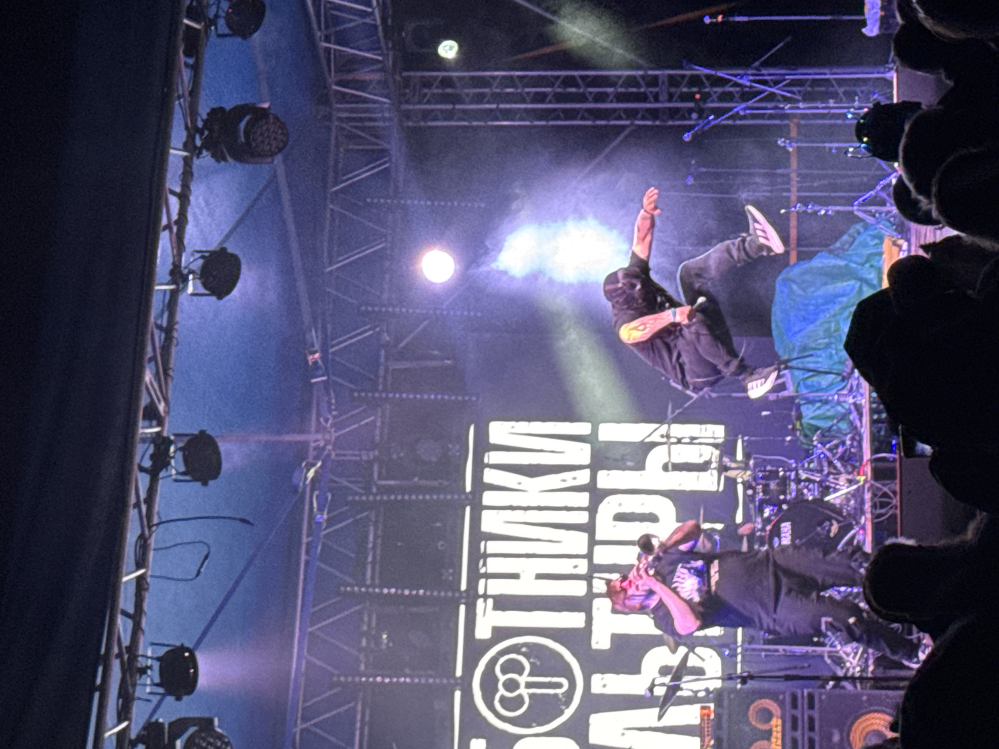 An outdoor concert stage in Большая Кивара, Russia, is depicted at night, illuminated by multiple stage lights casting blue and purple hues. A large white backdrop sign displaying Cyrillic text, reading "БОТНИКИ" and "ЛЬТУРЫ" (likely "РАБОТНИКИ КУЛЬТУРЫ" – "Workers of Culture"), is visible behind the performers. A stylized circular symbol, possibly representing intertwined 'P's or a treble clef, is on the left of the backdrop.
Two male performers are prominent on stage. On the left, a heavier-set man with reddish-brown hair and a beard, wearing a dark t-shirt and pants with white sneakers, is actively playing a brass instrument, likely a trumpet or cornet, held to his mouth. To his right, a second male performer, with long dark hair and wearing a black mask/balaclava, a dark t-shirt, dark pants, and black and white sneakers (with visible Adidas branding), is captured mid-jump. He is airborne, pushing off a blue draped object on the stage floor, with his left arm extended forward and his right arm bent. A distinctive orange flame-like design or tattoo is visible on his right bicep.
Behind the performers, a drum kit with "YAMAHA" branding on the bass drum is partially visible. Microphone stands are positioned on the stage. To the far left, large amplifiers or speaker cabinets are present. Overhead, metal trussing supports various stage lights, including circular LED fixtures and conventional spot/wash lights. A bright, circular white light source is visible above the jumping performer, with a vertical plume of light/smoke emanating from beneath it. The foreground shows the darkened heads of audience members, indicating a live performance setting.
