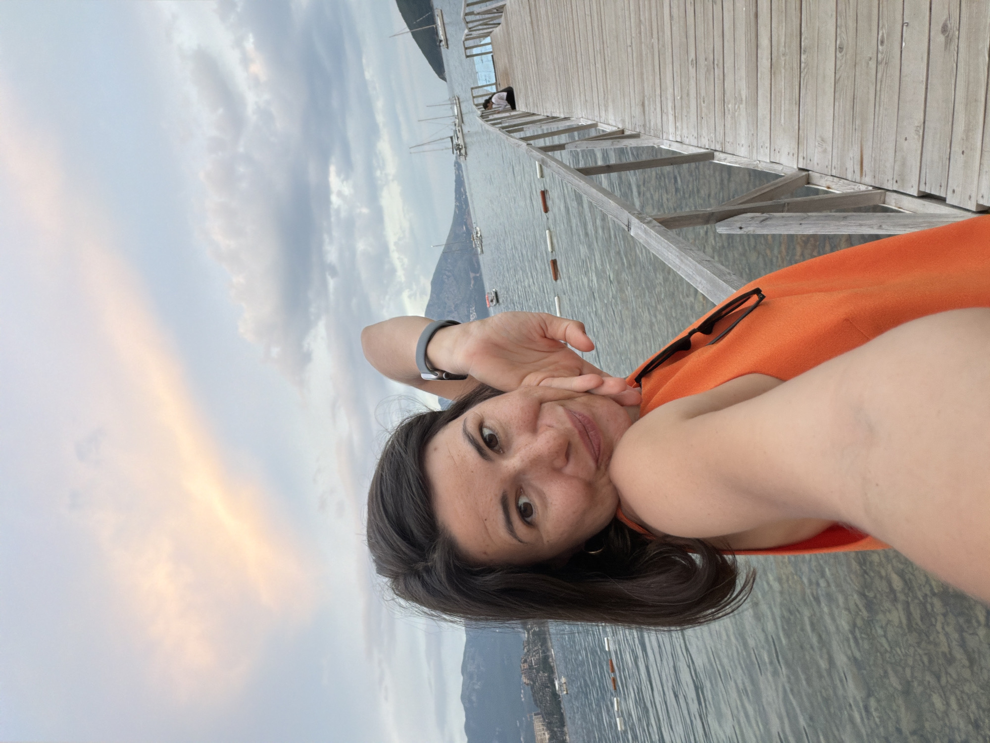 A woman takes a selfie on a wooden pier in Budva, Montenegro. The woman has dark hair and is wearing an orange top. She is smiling and has her hand on her cheek. In the background, there are sailboats on the water, mountains, and a cloudy sky with streaks of pink and orange from the sunset. The pier extends into the water, and there are buoys marking a swimming area. Another person is sitting on the pier in the distance.
