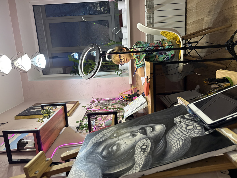 An indoor scene in a room located in Rostov-on-Don, Russia, likely a study or art studio, is captured at night. A young male child with light-colored hair and glasses, wearing a multi-colored short-sleeved shirt, sits at a wooden desk on a white plastic chair with a yellow cushion. He holds a pencil in his right hand, his left arm resting on an open notebook. His gaze is directed forward towards the viewer with a neutral expression.
To his right, a black tripod stand supports a large, illuminated LED ring light, positioned for bright frontal illumination. The desk, with black metal legs, also holds a red rectangular device, scissors, and other study materials.
In the left foreground, a wooden easel displays a large vertical canvas featuring a detailed monochrome drawing of a woman's face with vacant white eyes and cracked skin, intertwined with two snakes, one near her face and another larger one at the bottom. Below the canvas, on the easel's lower shelf, a white tablet in a black case lies beside a holder filled with art brushes and pens.
The room's walls are painted a light pink. The left wall features a black-framed wooden shelf holding a clear cylindrical object and a small potted plant. Below it, a black wire grid panel is adorned with artificial green ivy, pink ribbons, and small photographs. Another framed artwork with a gold frame is visible further up on the wall.
A large window on the right side of the room reveals a dark, indistinct exterior, suggesting evening or night. The windowsill holds several small white potted plants. Below the window is a white radiator. Three stacked, geometric pendant lights illuminate the ceiling. The floor is wooden.