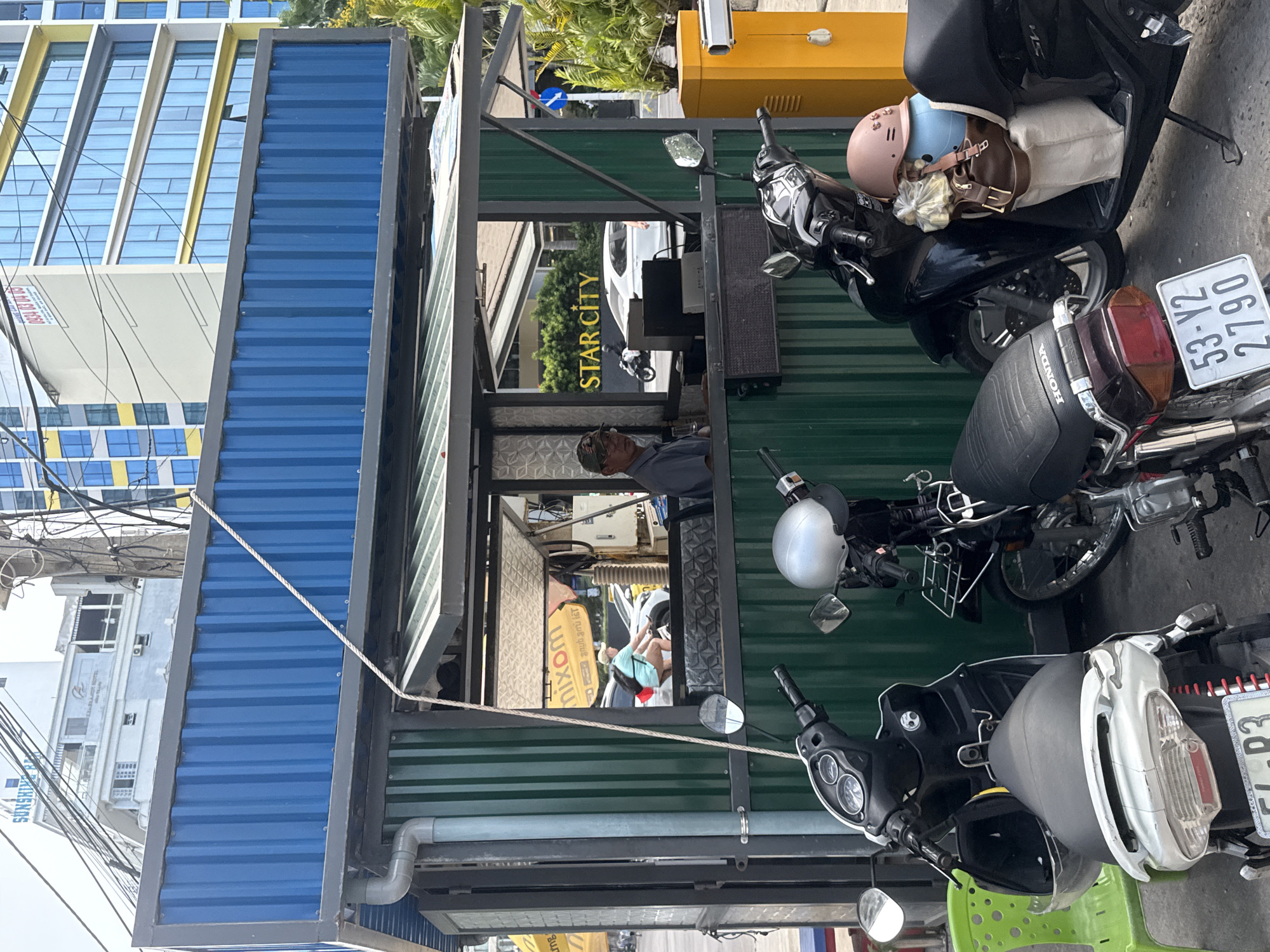 This image captures a vibrant urban street scene in Nha Trang, Vietnam, during clear daylight hours. In the foreground, several motorcycles are parked in front of a small, two-part structure. The main part of the structure is a kiosk or booth made of green corrugated metal panels, featuring an open window or serving hatch. Inside, a man wearing a cap is visible, looking out. Another person in a light blue top is faintly discernible further inside, possibly a reflection or another individual. Above and to the left of this green section, an upper structure made of blue corrugated metal panels extends upwards.
Numerous motorcycles are parked beside the green structure. One black scooter carries a pink helmet, a light blue helmet, a brown leather bag, and a plastic bag filled with items on its seat. Another scooter has a silver helmet on its handlebars. License plates are visible on some vehicles, including "53-Y2 2790" and "54-P3", consistent with Vietnamese registration. The word "HONDA" is embossed on the seat of one scooter.
In the background, a tall, modern building with a facade of blue windows and yellow frames dominates the left side of the frame. Further down the street, a white car is visible, and the words "STAR CITY" are prominently displayed on a building. A yellow barrier or machine stands near the parked scooters. Electrical wires crisscross the sky in the upper left, and a white rope or cable stretches across the middle of the green structure. A grey pipe is attached to the bottom of the green section. A small portion of a green plastic chair is visible in the lower right. The setting appears to be a busy commercial area.