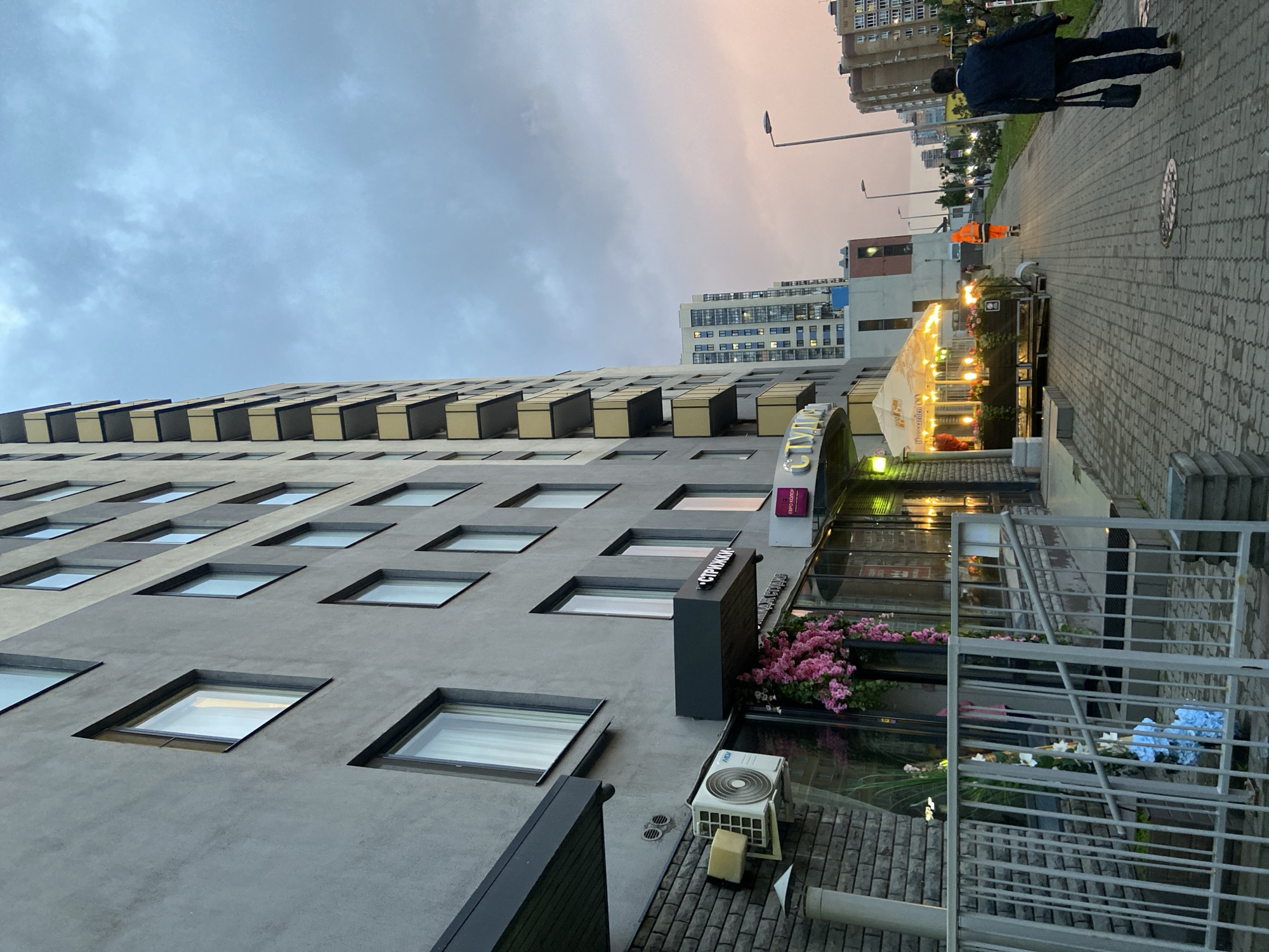 This media file captures an urban street scene at dusk or early evening. The prominent subject is a modern, multi-story building with a grey facade and numerous rectangular windows. The upper levels of the building feature distinct, recessed rectangular elements that create a geometric pattern.
At the ground level of this building, there is a commercial establishment, likely a cafe or restaurant, with large glass windows reflecting the surroundings. An outdoor seating area is visible under a white tent illuminated by string lights. Planter boxes filled with pink flowers decorate the entrance, and another arrangement of blue flowers is seen behind a metal railing. An air conditioning unit, bearing the brand "Midea," is mounted on the wall near the entrance.
A paved sidewalk runs along the right side of the image, where two people are walking into the distance. One individual is dressed in a dark suit, carrying a bag, while the other wears a bright orange safety vest. Streetlights are visible along the sidewalk, and a manhole cover is embedded in the pavement. In the background, other residential or commercial buildings can be seen, hinting at a developed urban environment.
The sky transitions from a light blue with scattered clouds on the left to a warm pink and orange hue on the right, indicative of a sunset. Artificial lights from the tent, streetlights, and some building windows are on, contrasting with the fading daylight.
Visible text includes "CITY" and "СТРИЖКИ" (Strizhki - Haircuts) on a main sign, "ЄВРО КУРОРТ" (YEVRO KURORT - Euro Resort) on a smaller sign above the entrance, and "НОВИНКА OD" (Novinka OD) on the tent.