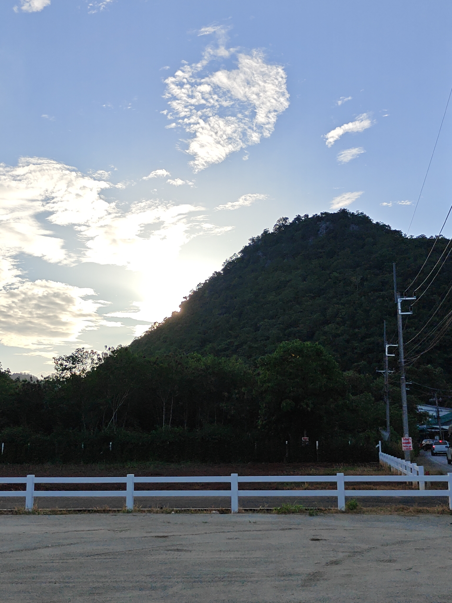 In the city of Mu Si, Thailand, this image captures a serene natural landscape under a partly cloudy sky. The sun, partially obscured by clouds and a steep, densely forested mountain slope, casts a warm glow. The mountain dominates the background, its dark green foliage contrasting with the lighter tones of the sky and the sun's rays. In the foreground, a white fence runs horizontally across the frame, separating a dirt or gravel area from a patch of grass and trees. Electrical poles and wires are visible on the right side of the image, suggesting the presence of nearby infrastructure. A few vehicles and buildings are faintly visible in the distance, also on the right, hinting at a residential or commercial area adjacent to the natural scenery. The overall atmosphere is tranquil, with the interplay of light and shadow on the mountain and clouds creating a dynamic visual texture.