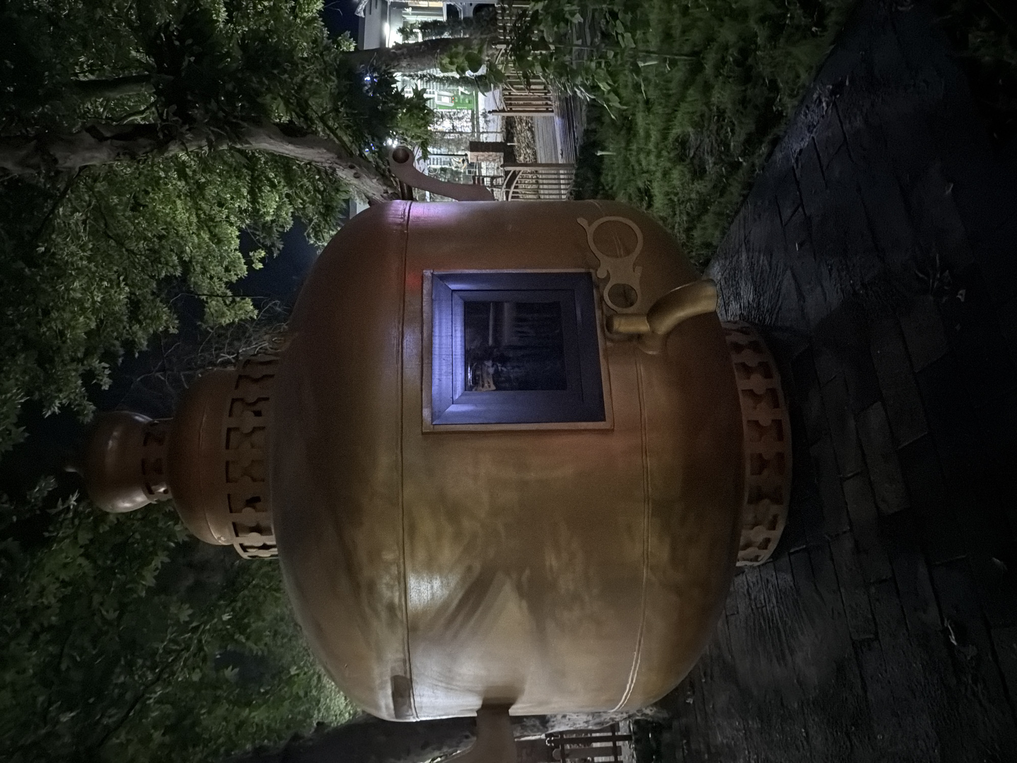 An outdoor nighttime shot in Chirchik, Uzbekistan, features a large, golden teapot-shaped structure that appears to be a kiosk or monument. The structure is illuminated, highlighting its metallic surface and intricate details, including a small window with a blue-lit interior and a spigot protruding from its side. The teapot is situated on a dark, paved area, possibly a walkway or plaza, which is wet and reflective, suggesting recent rain. Surrounding the structure is lush green grass and the shadowy forms of large trees with green foliage, illuminated by ambient lighting. In the background, a glimpse of a building with lights and a wooden fence can be seen, indicating a populated area. The overall atmosphere is somewhat mystical due to the low light and the unusual subject.