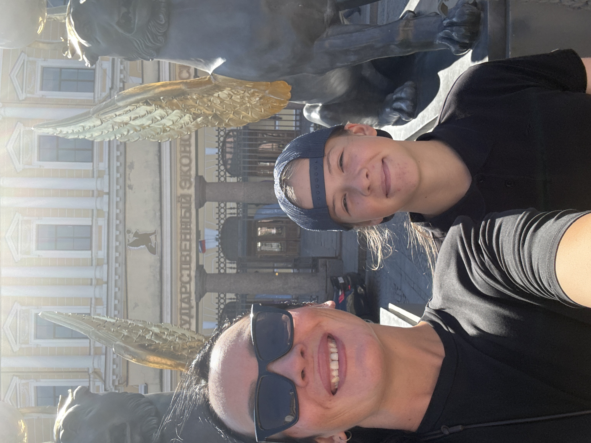 In Saint Petersburg, Russia, two individuals are captured in a selfie. The woman in the foreground, wearing sunglasses and a black t-shirt, is on the left. To her right is a younger person wearing a blue baseball cap and a black polo shirt. Behind them, a building with classical architecture, featuring columns and ornate windows, is visible. Two large, dark statues of winged griffins flank the building entrance. Gold wings are prominently featured on the griffins. The building has text above the entrance, which appears to read "ГОСУДАРСТВЕННЫЙ ЭКОНОМИЧЕСКИЙ". The overall scene suggests a tourist visit to a significant cultural or historical landmark in the city.