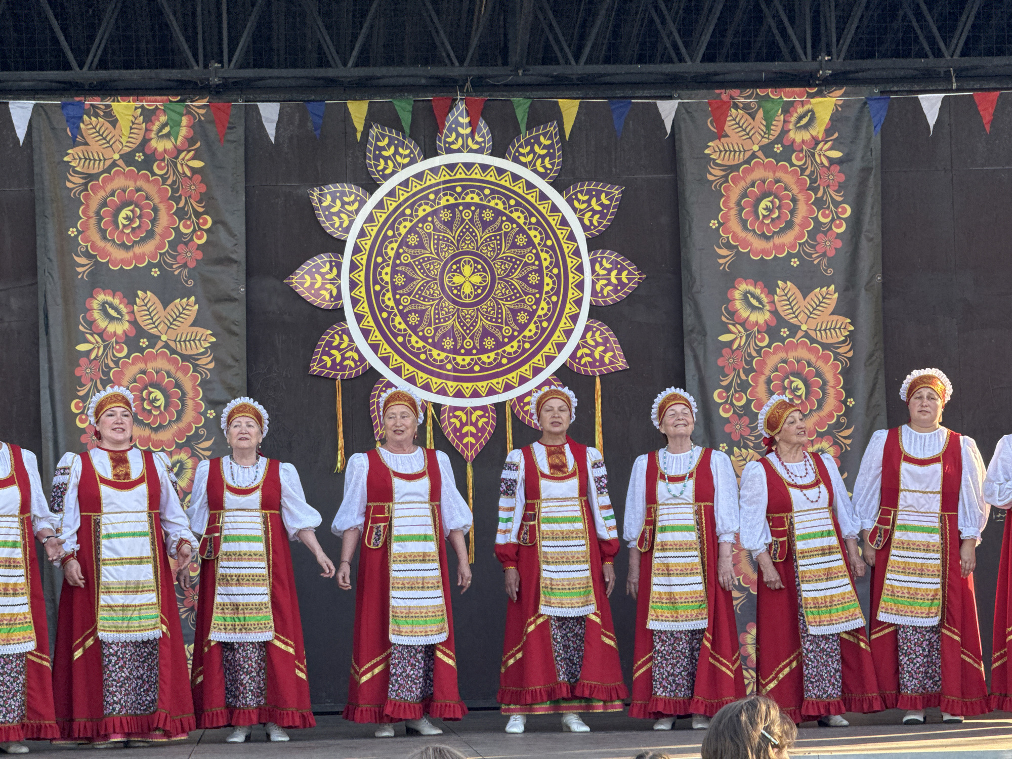 A group of women dressed in traditional Russian folk costumes are performing on a stage decorated with Russian-themed banners and a sun-shaped ornament. They stand shoulder-to-shoulder, holding hands, and facing forward. The banners in the background feature floral patterns, and a string of pennant flags hangs above them. The performance is taking place in Zav'yalovo, Russia.
