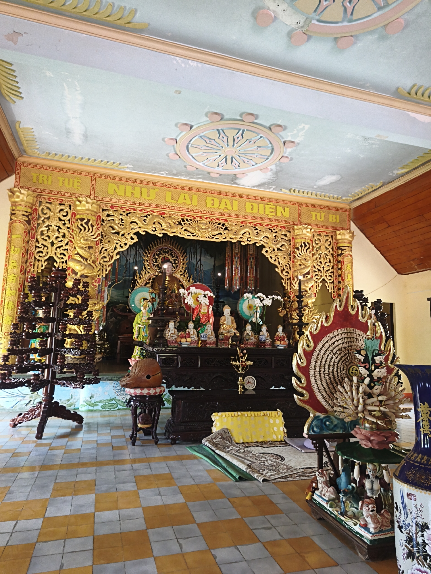 The image shows the interior of a Buddhist temple in Cam Ranh, Vietnam. The scene is dominated by a golden altar with various statues and religious artifacts. In the center, a large Buddha statue sits on a throne, flanked by smaller figures. To the left, a dark wooden tiered stand holds numerous small statues. In front of the altar, a patterned rug is laid out on a checkered floor. The ceiling is decorated with painted medallions and ornate trim. The overall impression is one of a richly decorated and sacred space.