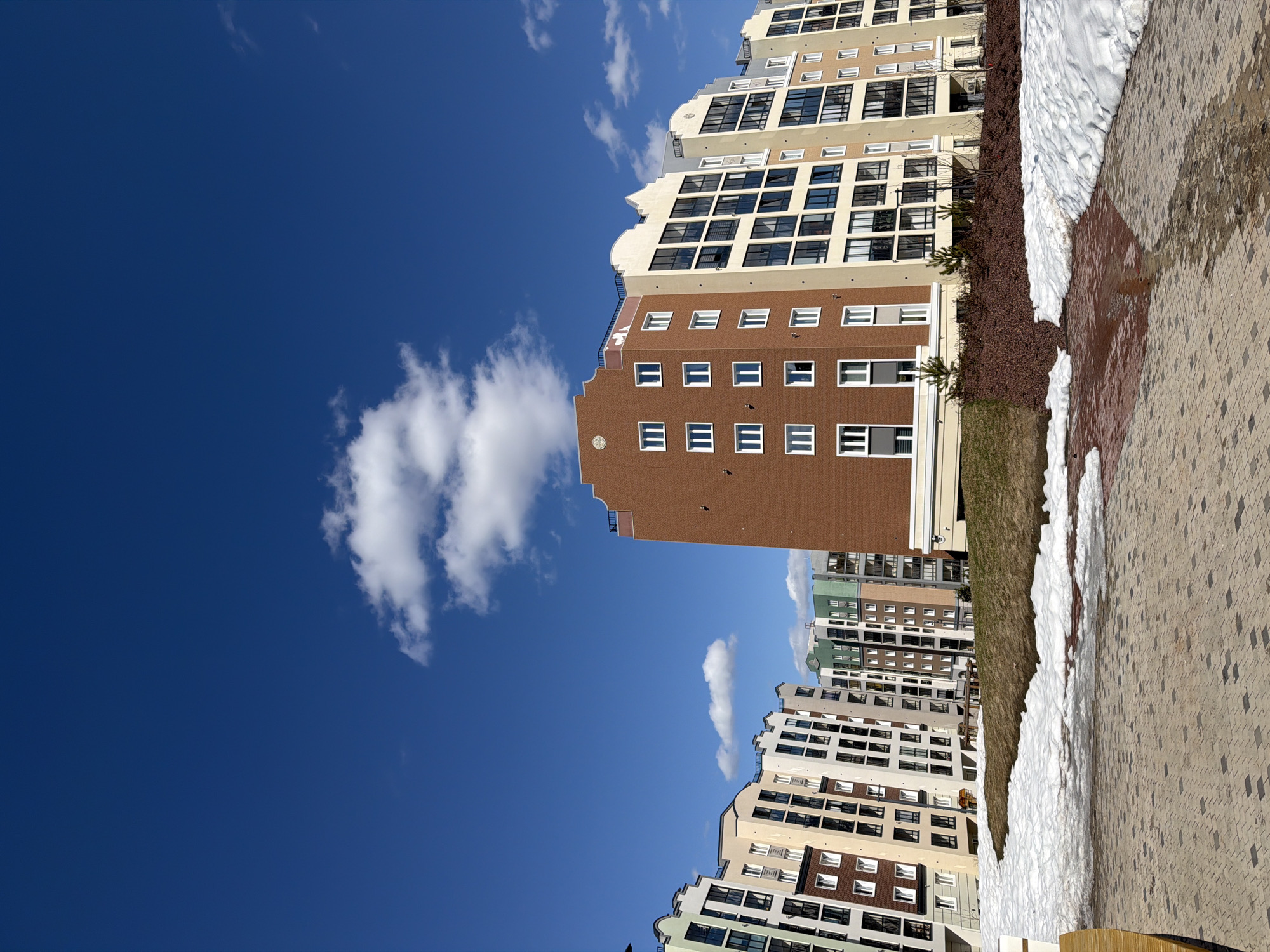 A brick building with rectangular windows is surrounded by other buildings with similar architectural styles against a bright blue sky with scattered white clouds. The buildings are located near snow-covered areas. The location is in the city of Pervomaysky, Russia.
