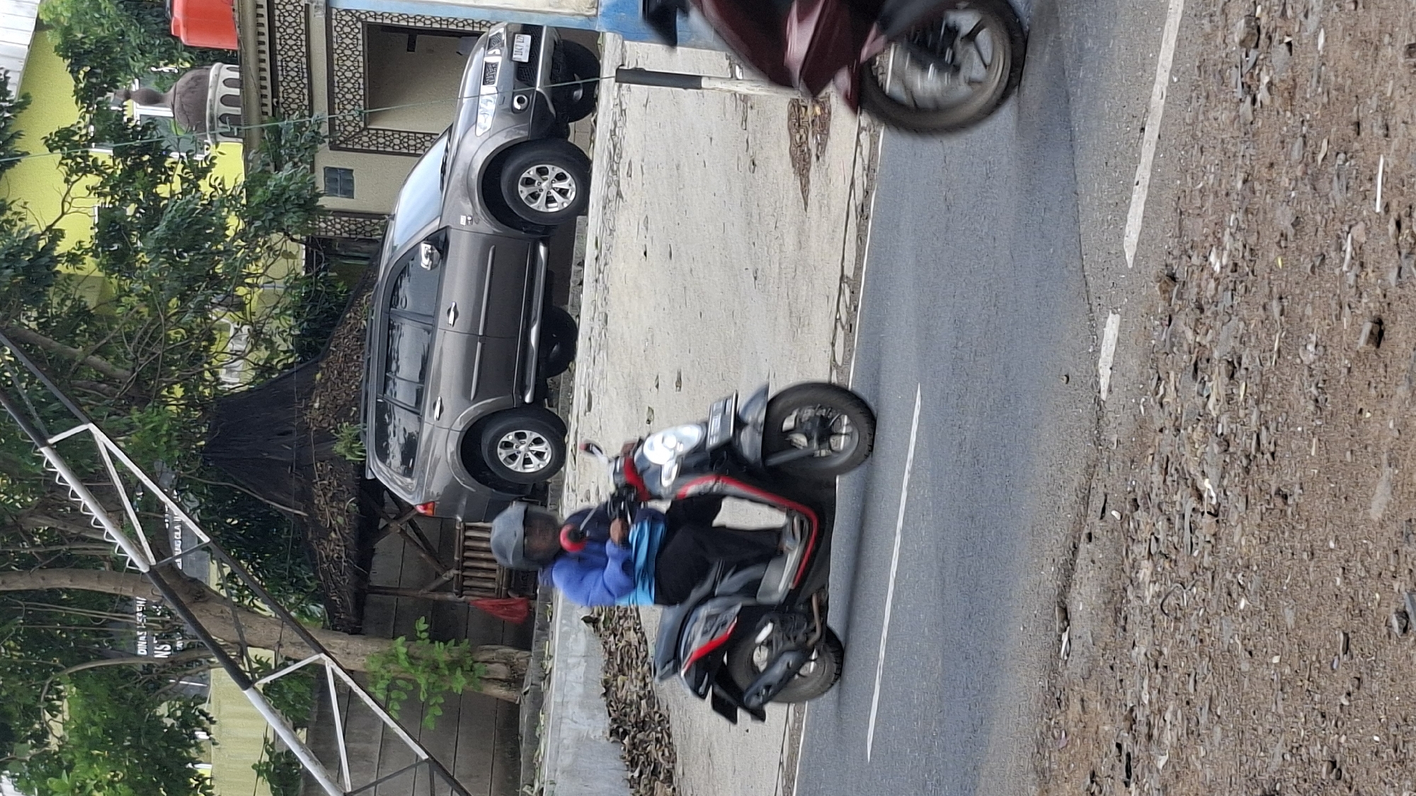 The image depicts a street scene in Cilaku, Indonesia, with a paved road in the foreground. A person is riding a black and red scooter, wearing a blue jacket and a black helmet. Behind the scooter, a gray SUV is parked on an elevated area next to a building with a green wall and a dome. Trees with dense foliage are visible in the background, along with what appears to be a thatched-roof structure. The scene suggests a typical day with traffic and urban elements.