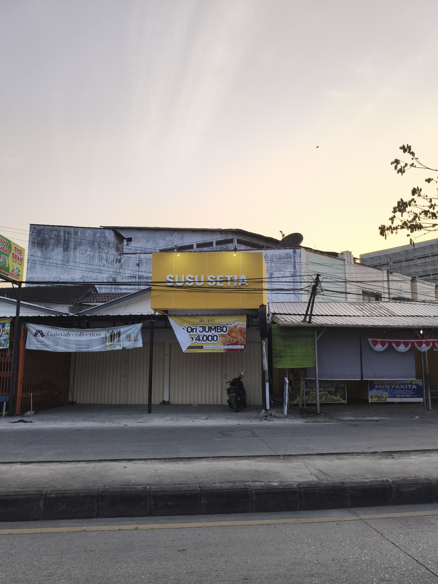 The image shows a street scene in Samarinda, Indonesia, likely in the afternoon or early evening given the warm light and the sky's gradient. The foreground features a street with a concrete curb and asphalt pavement. In the middle ground, there's a row of businesses. A prominent yellow sign with the text "SUSU SETIA" is mounted above a building. Below this, several storefronts are visible, some with corrugated metal roll-up doors. Banners are hung in front of some of these establishments. One banner reads "Tiaisya collection" and displays images of clothing and accessories. Another banner advertises "Ori Jumbo 14,000". Further down the row, there are signs for "MINYAKITA" which is a brand of cooking oil. A parked motorcycle is visible in front of one of the shops. The background consists of buildings of varying heights and architectural styles, with power lines and a satellite dish also present. Trees with foliage are visible on the right side of the frame, and a bird can be seen in the sky on the upper right. The overall impression is one of a commercial street with an active, albeit subdued, atmosphere.
