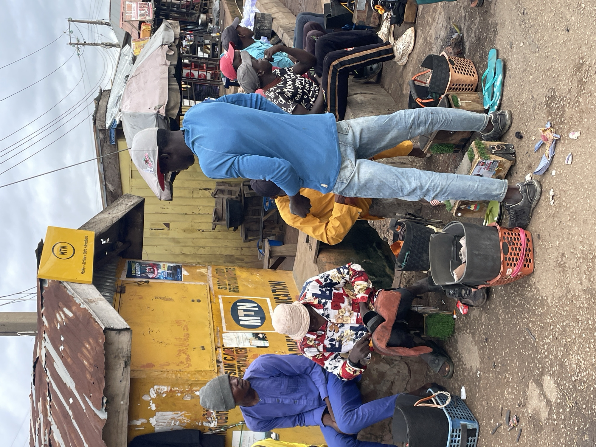 In Jos, Nigeria, this image captures a bustling street scene. A vibrant yellow structure, likely a kiosk, dominates the foreground, adorned with a prominent "MTN" sign, indicating a telecommunications service provider. A group of individuals are gathered, some seated on a low wall or ledge, while others stand.
One man, dressed in a blue long-sleeved shirt and blue trousers, sits on the left. Next to him, another man in a colorful, patterned shirt and a light-colored bucket hat is engaged in an activity, possibly repairing or polishing an item held in his hands. To the right, a young man wearing a blue long-sleeved shirt, light-wash jeans, and a baseball cap stands facing away from the camera, looking towards the street. He appears to be conversing or observing. Further to the right, several other individuals are seated, some with heads bowed or looking towards the left.
Various objects are scattered around the scene. Baskets, some containing items, are placed on the ground. Shoes, including sandals and sneakers, are visible. Packaging materials and debris litter the ground, suggesting a commercial or market environment. The background reveals more stalls or shops with corrugated metal roofs, and overhead wires crisscross the sky, typical of an urban setting. The overall atmosphere appears to be one of everyday commerce and social interaction in this Nigerian city.
