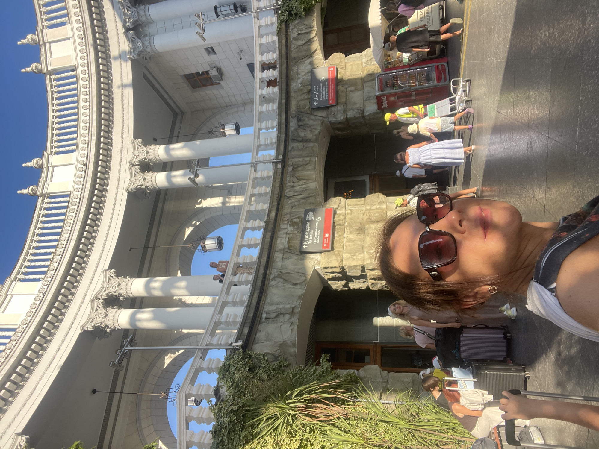 A woman is taking a selfie in front of a grand building with white columns. The building appears to be an architectural landmark with a curved facade and intricate detailing. Several other people are visible in the background, some with luggage, suggesting a travel hub like a train station or airport.
In the background, signs are visible, one of which reads "К поездам" (To trains) with an arrow pointing left, indicating a direction to train platforms. Another sign with "Доброй" is also partially visible. The weather appears sunny with clear blue skies. The time of day is likely daytime, given the bright sunlight casting shadows. The woman in the foreground is wearing sunglasses and a patterned top, her expression is relaxed. The overall scene conveys a sense of travel and vacation. The architecture suggests a location in Sochi, Russia, possibly a train station given the signage and luggage.