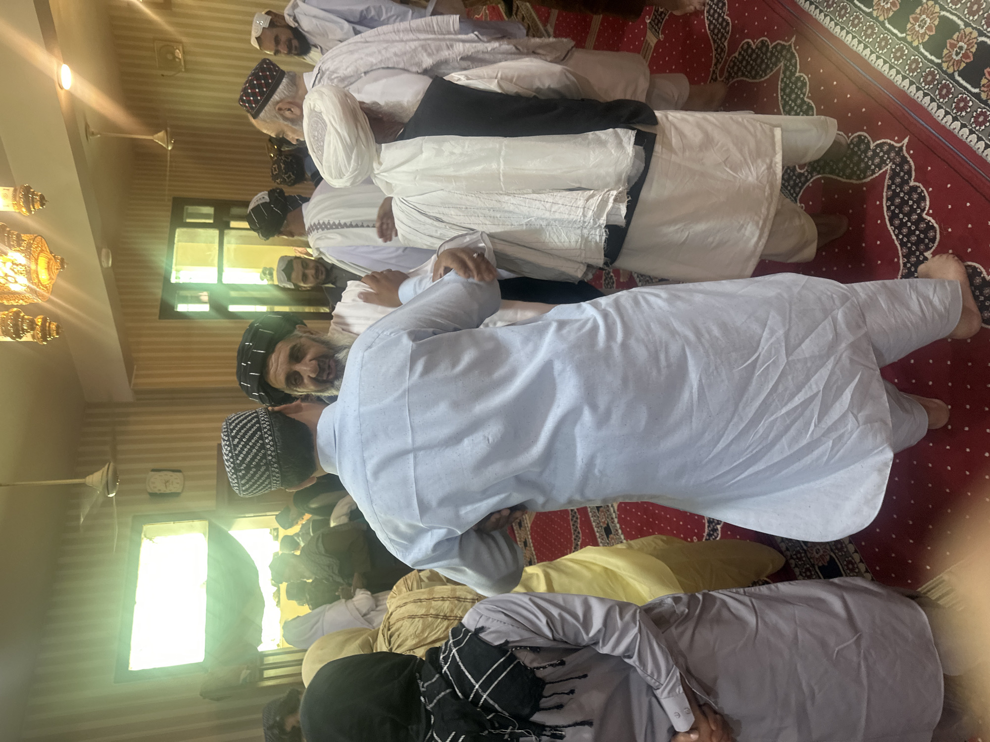 A group of men are praying in what appears to be a mosque in Quetta City Tehsil, Pakistan. They are wearing traditional Muslim clothing. The room has a red patterned carpet and a chandelier. Some of the men are wearing distinctive hats and turbans. The photo is taken from an overhead perspective.