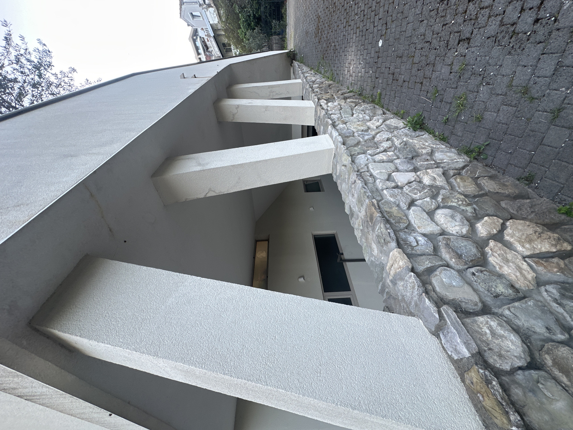 The image presents an architectural exterior scene, viewed from a low, upward-looking perspective, likely an entrance or side of a building. A modern structure, characterized by its clean lines and white, textured facade, dominates the left side. Several prominent, rectangular support beams, also white and textured, extend horizontally from the building, creating an overhang.
Adjacent to the building on the right is a sturdy retaining wall constructed from irregular, grey stones cemented together. This stone wall separates the building area from a dark grey paved surface, which appears to be a driveway or pathway, made of interlocking paver blocks. Small patches of green grass and weeds are visible growing between the cracks of the pavers and along the base of the stone wall.
In the upper left quadrant, a tree with sparse, dark branches is silhouetted against a bright, overcast sky. Parts of other buildings, possibly residential, with traditional architectural elements, can be seen in the distant background, hinting at an urban or suburban setting.
The scene depicts no human activity or specific event, conveying a sense of stillness. The bright, diffused light suggests it is daytime, possibly late morning or early afternoon, under cloudy conditions. No discernible text is visible anywhere in the image. The architectural style and materials, including the stone wall and paver blocks, align with common aesthetics found in locations such as Budva, Montenegro.