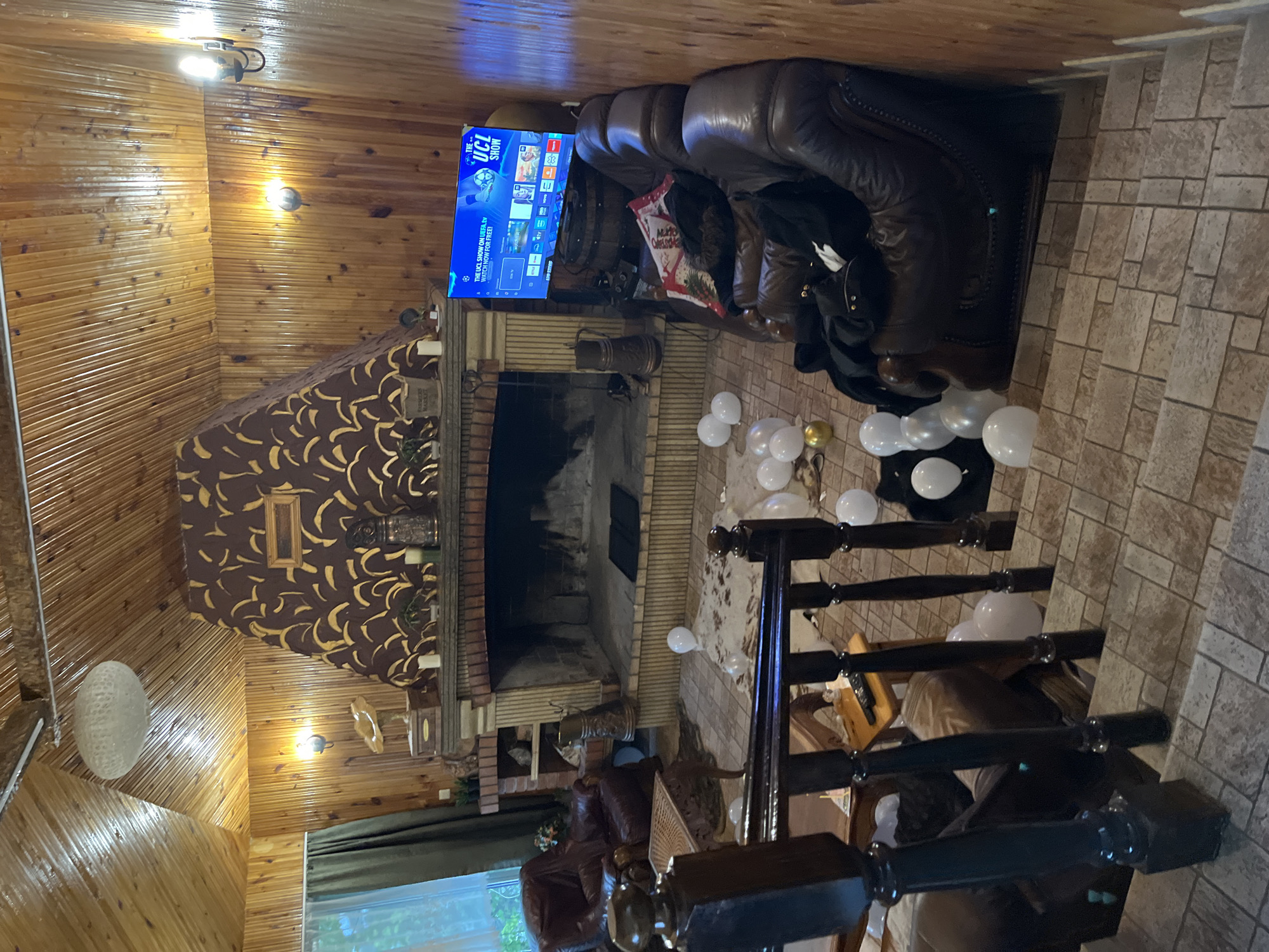 The image shows the interior of a room, likely in a house or cabin, possibly located in or near Presoka, Bulgaria, given the user's prompt. The room features wooden paneling and beams, a large fireplace with a decorative surround, a television mounted above the fireplace, and a large leather sofa. White and gold balloons are scattered on the floor near the sofa. A staircase with dark wood railings is visible in the foreground. The environment appears to be a cozy, rustic living space, possibly a vacation home or lodge. There are no people visible in the image.
