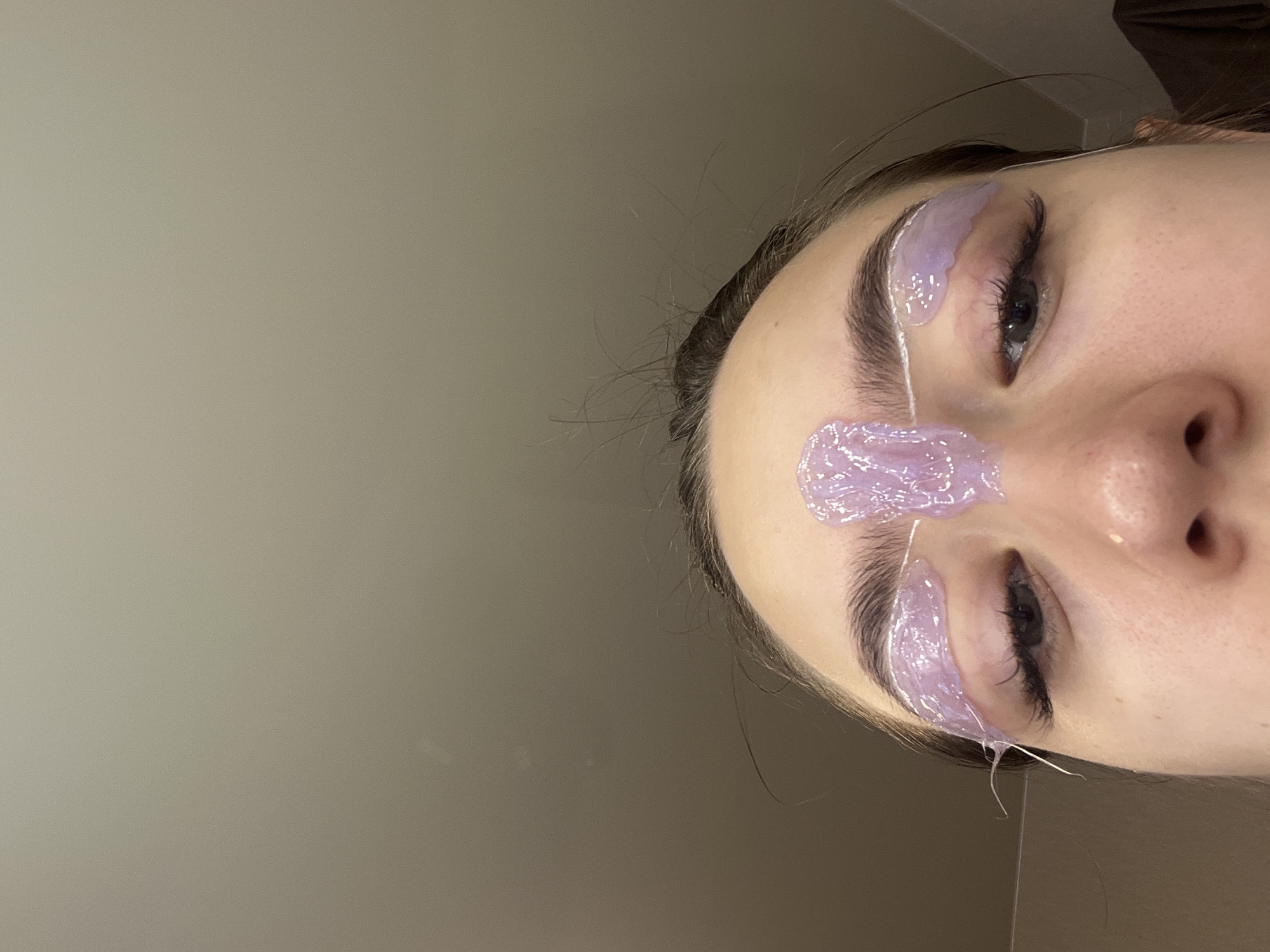The image is a close-up, low-angle shot of a young woman's face, focusing on her eyes and eyebrows. She has dark hair and appears to be undergoing some form of beauty treatment. Purple gel, possibly a mask or wax, is applied in strips above and between her eyebrows, with fine white threads also visible in the same areas. Her eyebrows are well-defined, and she has long eyelashes. The background is a plain, lightly colored wall, with some indistinct objects or textures visible on the right side of the frame. The lighting is soft and diffused. The image does not provide any specific information that would allow for its location to be identified within the city of Chaikovsky, Russia, or any other specific geographic location.