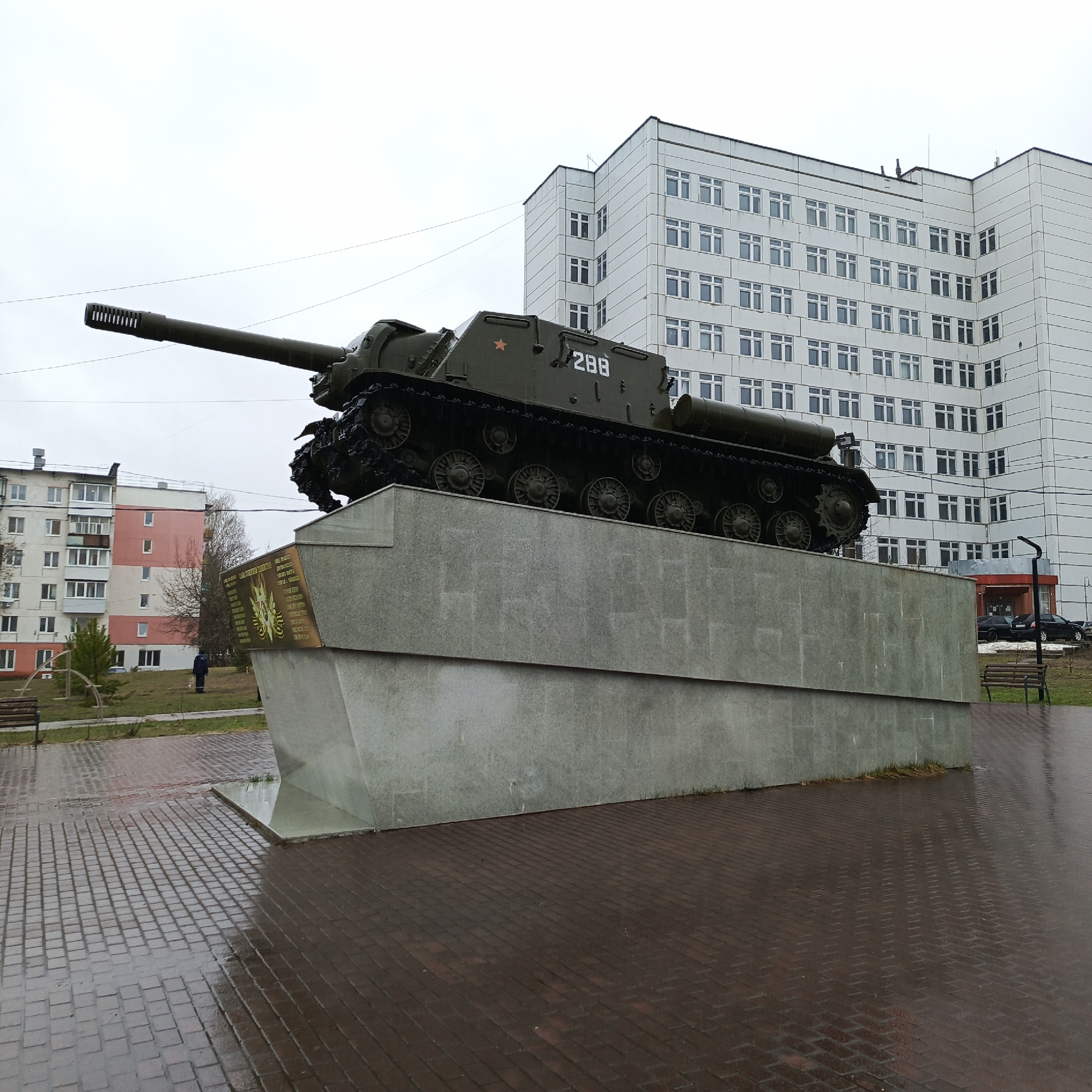 This image depicts a monument featuring a Soviet-era self-propelled gun, likely an ISU-152, set in a public square or park in Chaikovsky, Russia. The dark green military vehicle rests on a large, multi-tiered concrete pedestal, which is grey and angled. A red star insignia is visible on the side of the tank, along with the number "288" painted in white. The tank's long barrel extends towards the left, with a muzzle brake featuring multiple vents.
On the front of the pedestal, beneath the tank, there is a bronze-colored plaque with inscribed text, which is too small to read but appears to list names or commemorate an event.
The weather appears overcast and damp, with a grey sky and visible moisture on the paved ground, suggesting recent rain. The ground leading up to the monument is made of reddish-brown paving stones.
In the background, two prominent buildings are visible. To the right of the tank, a large, modern-looking multi-story building with numerous small, evenly spaced windows and a light-colored, possibly white or pale grey, facade stands tall. To the left, behind the monument, a residential apartment building with several stories, balconies, and a combination of white and pink-painted sections can be seen. A solitary person, dressed in a dark blue jacket, walks on the grass between the monument and the residential building in the distance. Benches are sparsely placed in the area, and some bare trees and sparse greenery are also visible. The scene captures a static monument in an urban public space on a cloudy day.